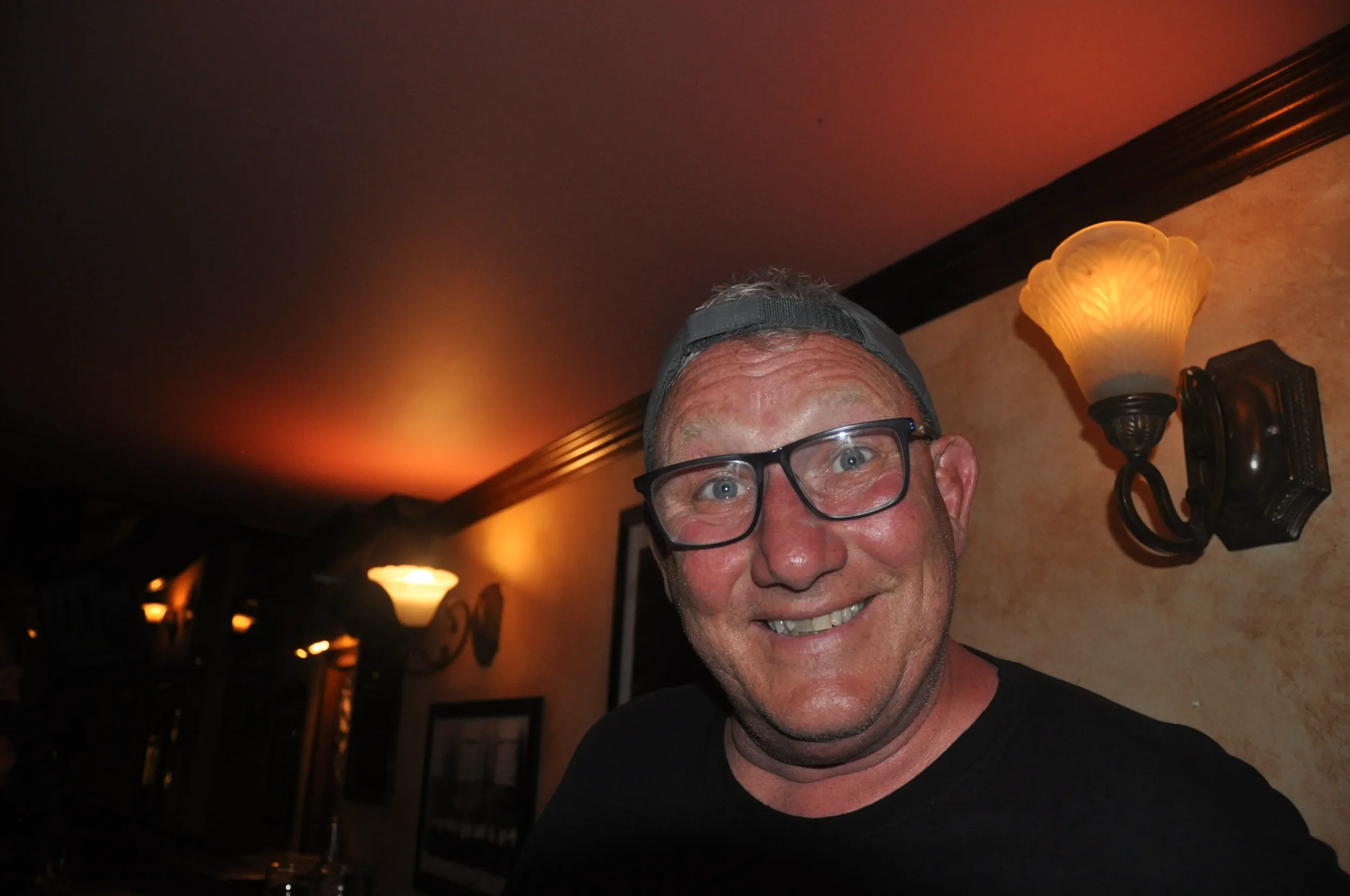 A smiling man with glasses and a backwards cap in a dimly lit room with vintage wall sconces.