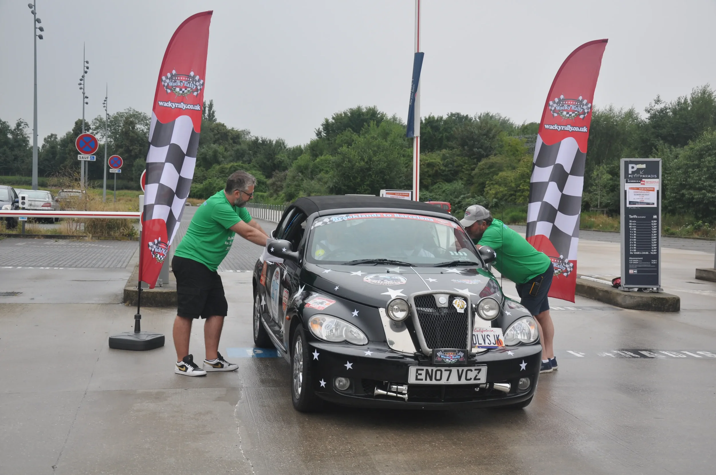 A black rally car with star decals being pushed by two men in green shirts at a rally event. The car has a California license plate 'EN07 VCZ' and is near red rally flags with checkered pattern and a parking sign in the background.