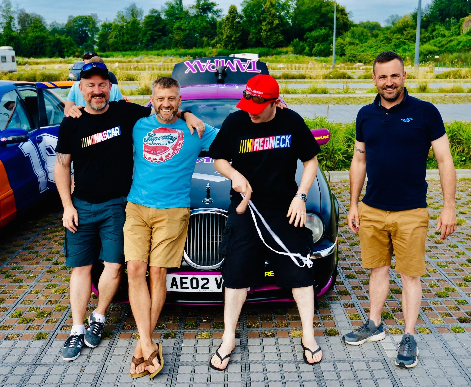 Four men stand in front of a black racing car with racing stripes, smiling for a photo outdoors on a sunny day. Two are wearing shorts, one has a red cap, and all are dressed casually.