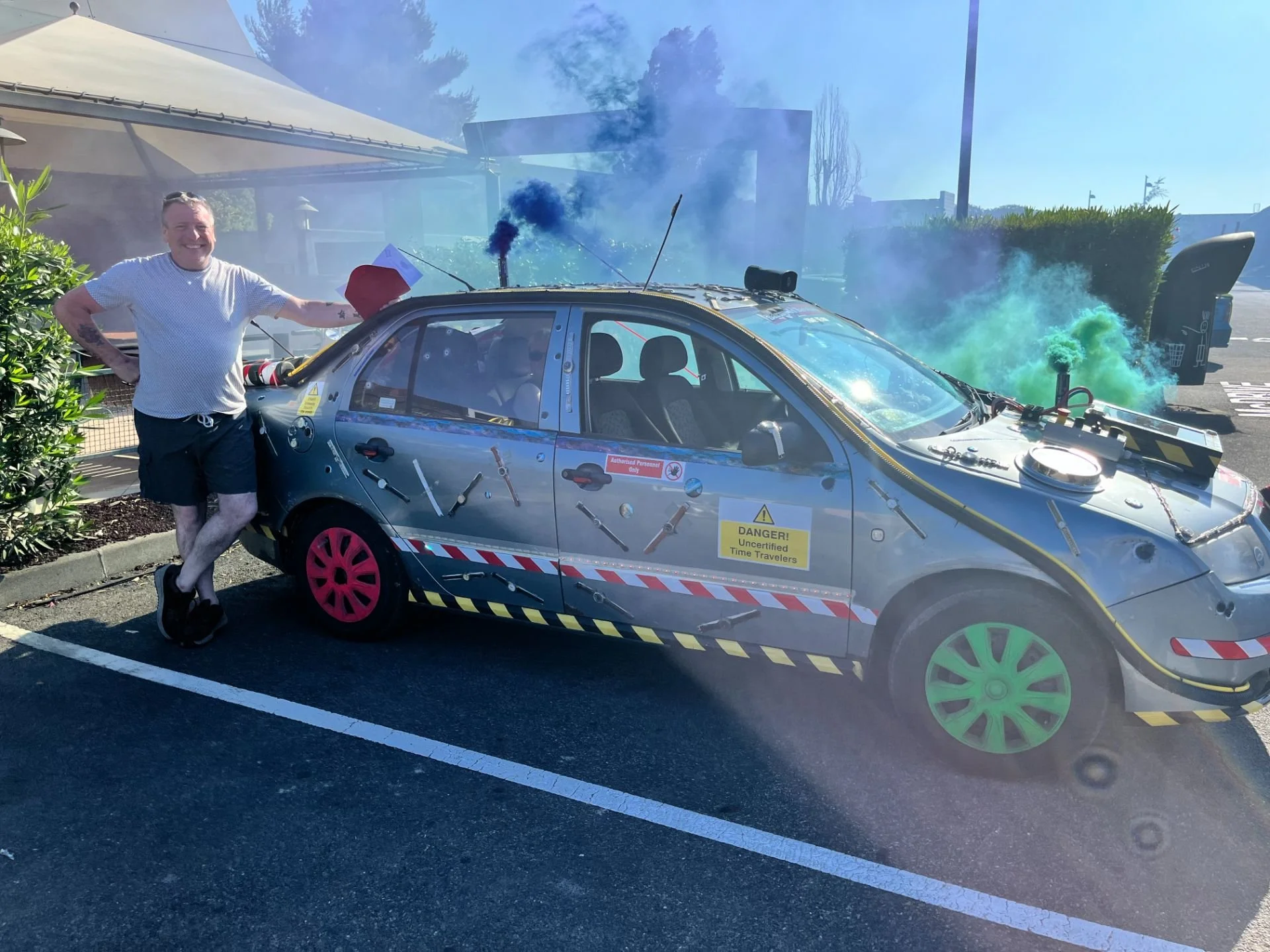 A man standing next to a Saskatoon police decorated car with colorful wheels, smoke, and fog bombs in the background, on a sunny day.