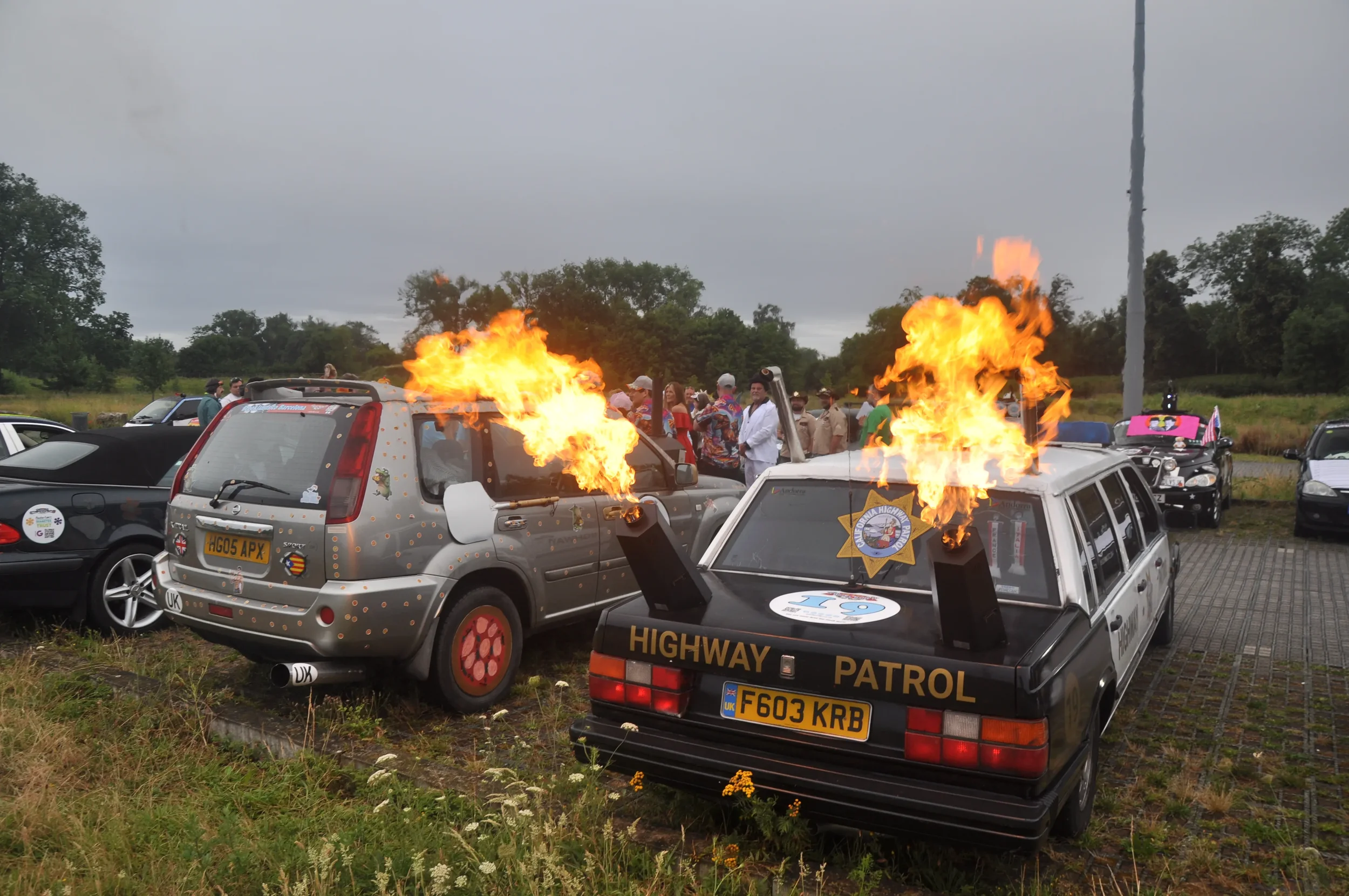 Cars with flaming exhaust pipes parked outdoors during a gathering, with people in the background.