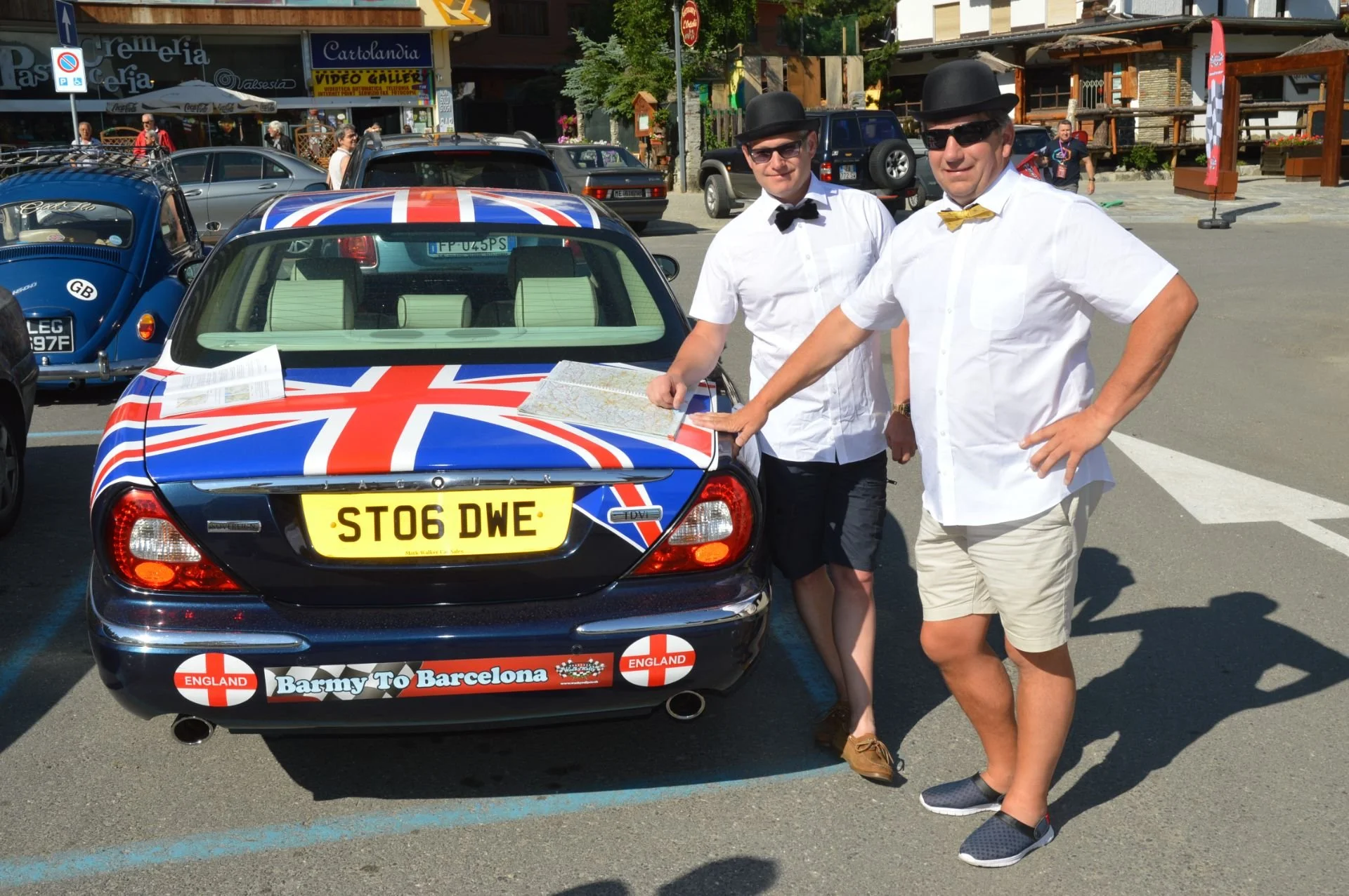 Two men in white shirts, shorts, and bow ties standing next to a car with a Union Jack flag design, a yellow license plate reading 'ST06 DWE', and stickers that say 'England' and 'Barmy To Barcelona.' One man is pointing to a map on the car, and the 