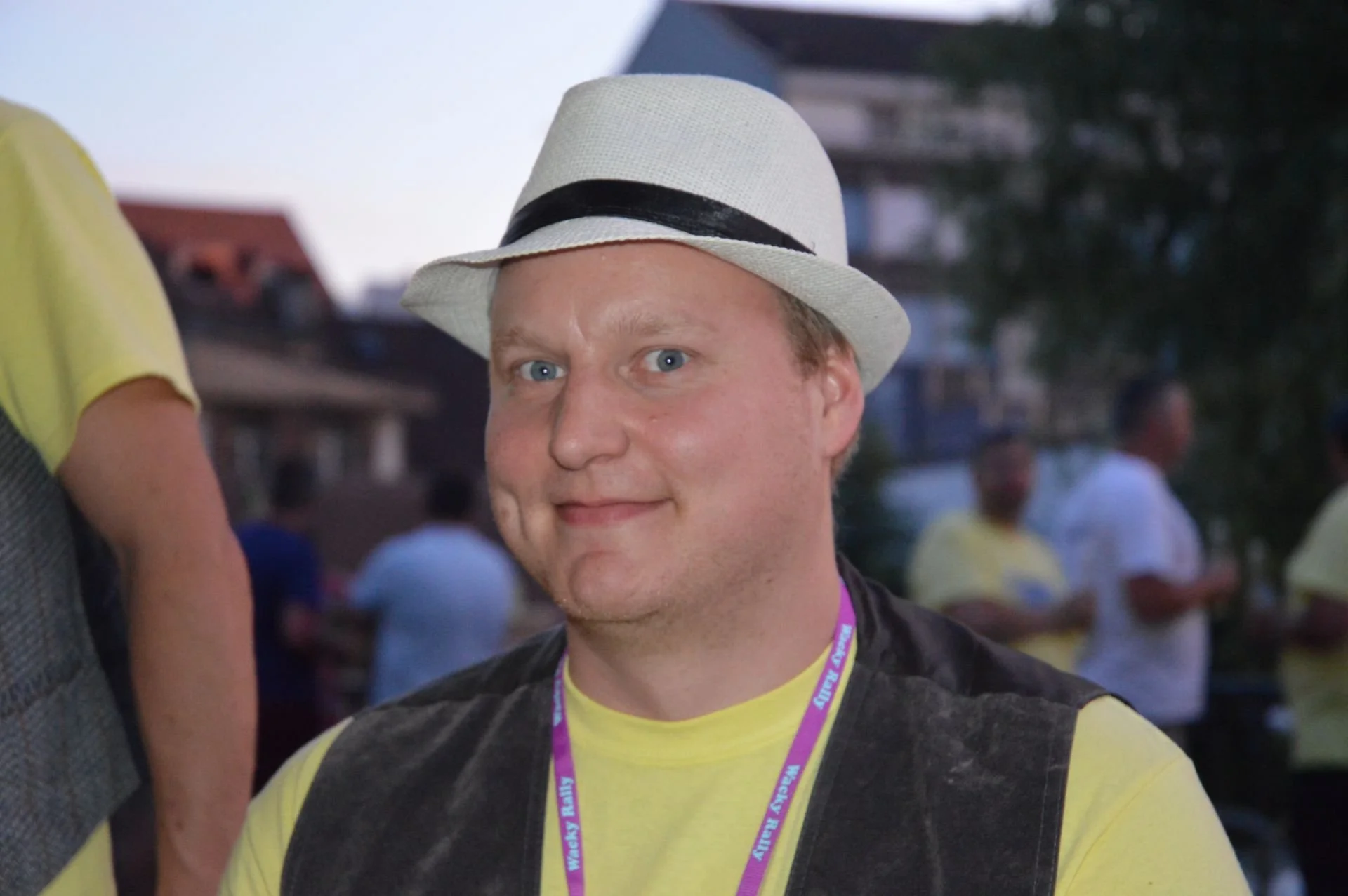 Person wearing a white fedora hat with a black band, a yellow T-shirt, and a black vest, smiling at the camera during an outdoor event with several people in the background.