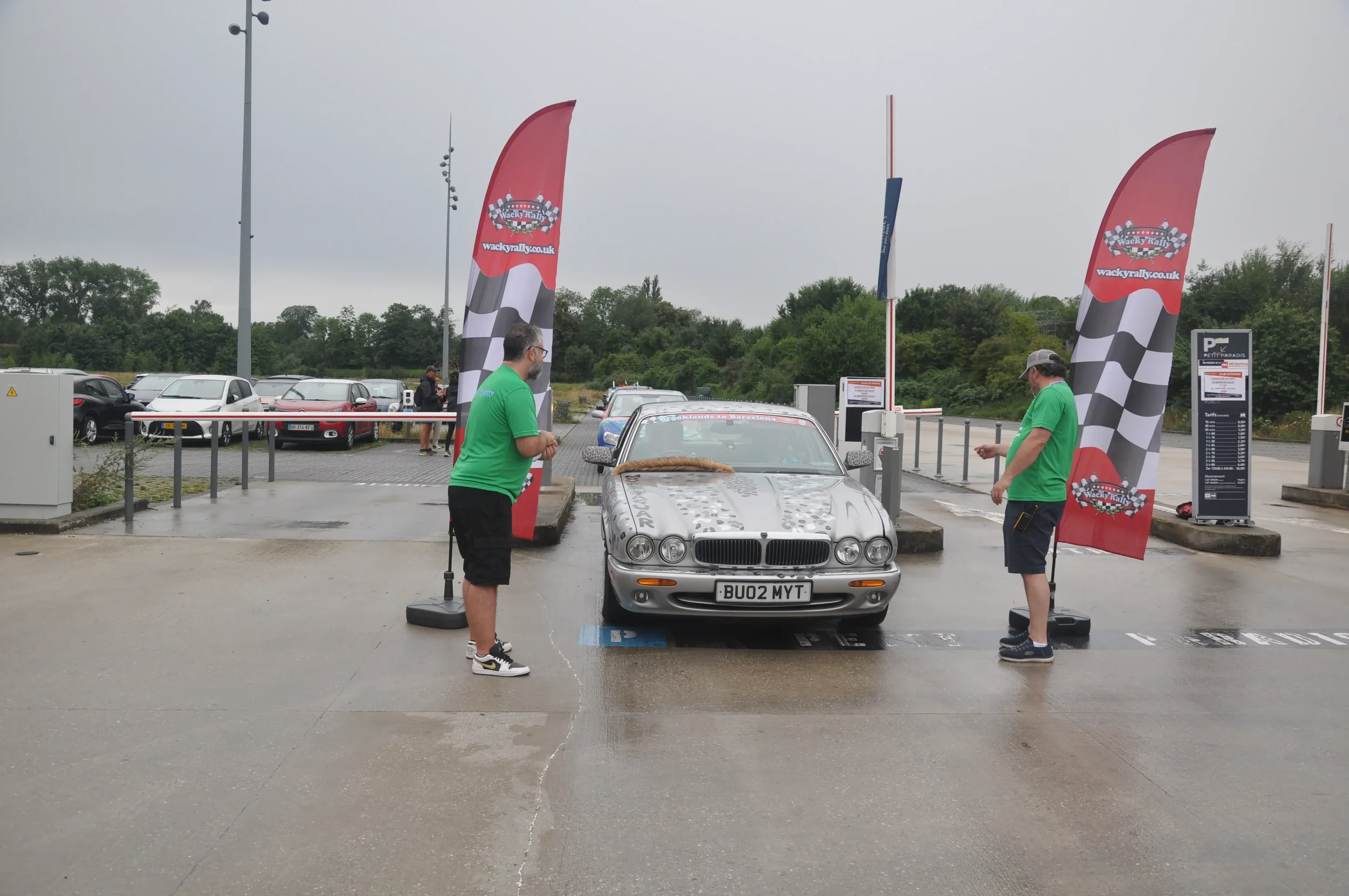 Two men in green shirts are standing on either side of a silver car in a parking lot with Wacky Rallies banners, overcast sky, and a line of parked cars in the background.