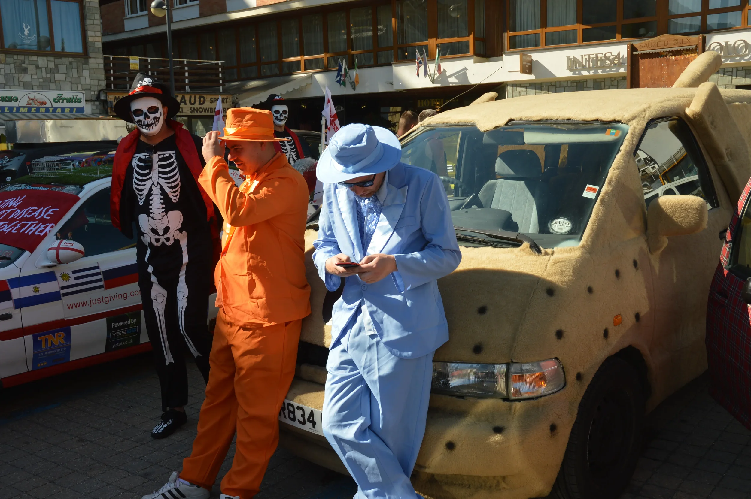 People in costumes stand beside a cardboard car and a decorated event vehicle in a street parade.