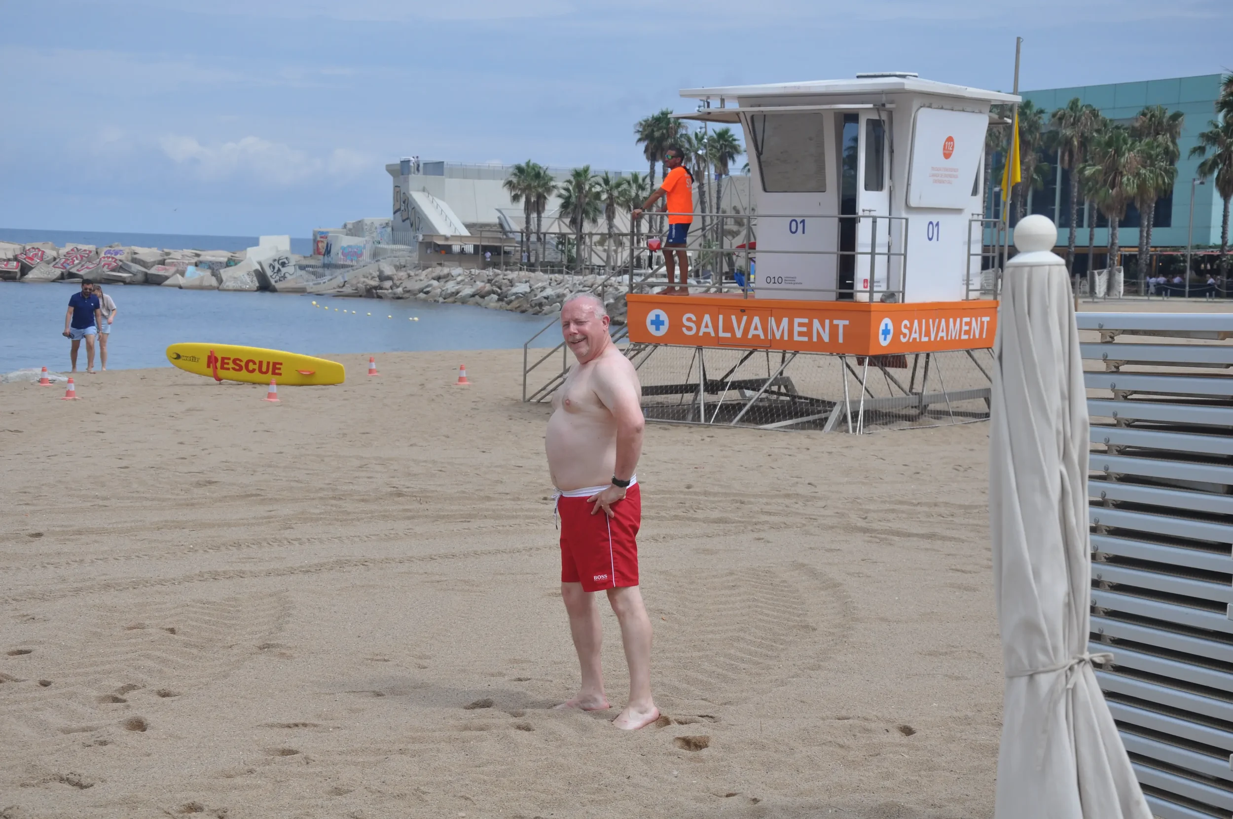 A man in red swim trunks standing barefoot on the sandy beach, smiling at the camera near a lifeguard station. The station is marked with orange and blue signage and has a lifeguard watching over the beach. A rescue board is on the sand nearby, and a