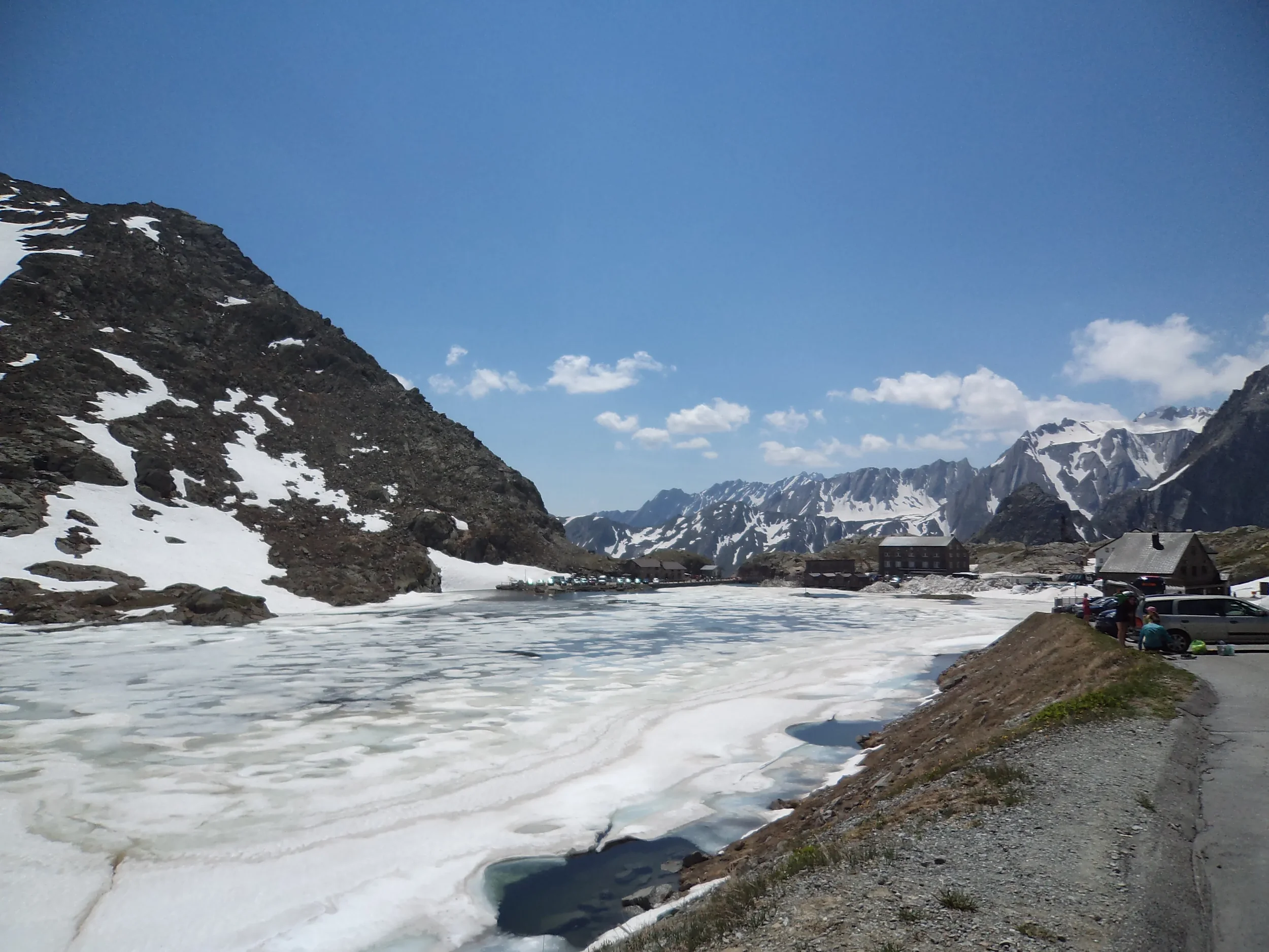Snow-covered mountains and a partially frozen lake under a blue sky with clouds, with parked cars and buildings along a roadside.