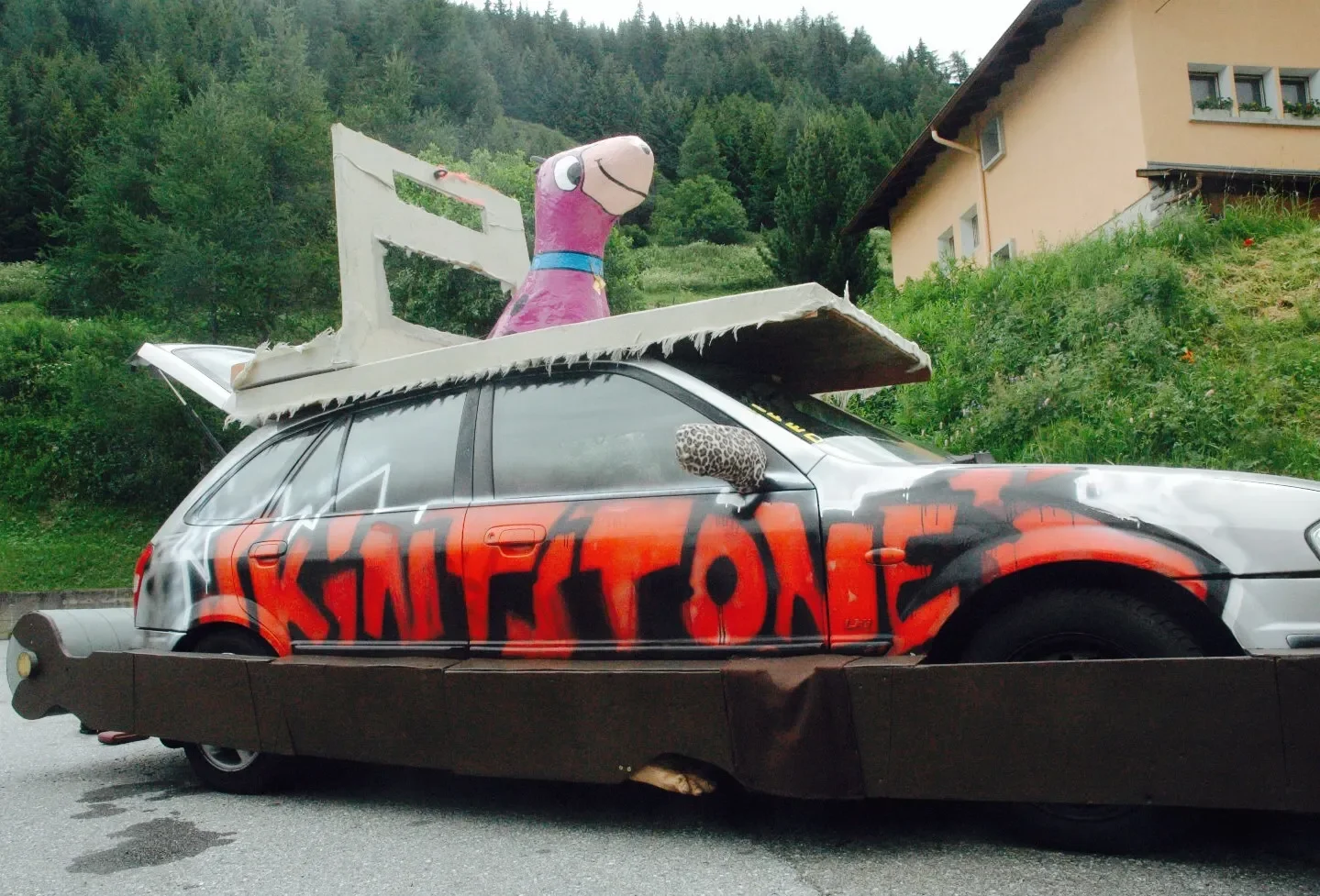A decorated car with graffiti reading 'SKIFETONE' on its side, topped with a large cartoon dog figure on a wooden platform, and featuring a leopard-print side mirror, parked on a street with a green hillside and a house in the background.