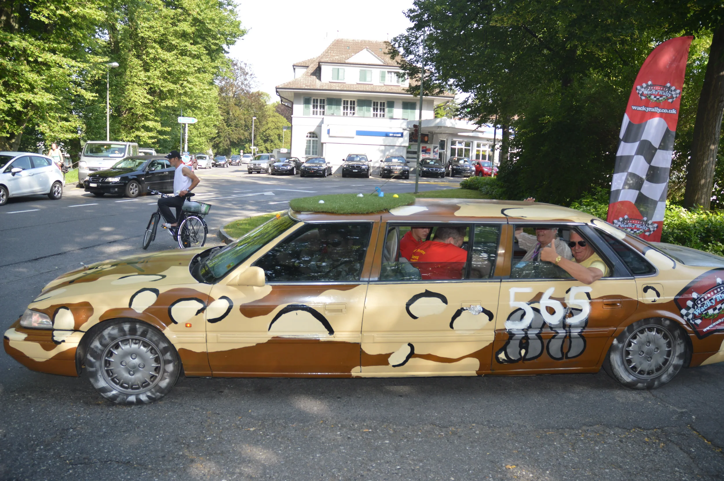A car decorated with a camouflage pattern and a grass patch on top, parked on a street. Several people are inside and around the car. A person on a bicycle is nearby, and there is a red banner with a checkered flag design in the background.