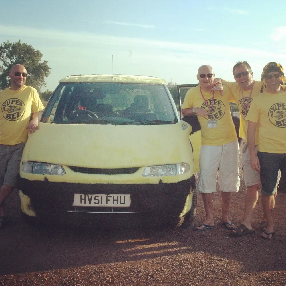 Group of men wearing yellow 'Super Bee' T-shirts posing next to a yellow and black vehicle with a custom front design resembling a bee.