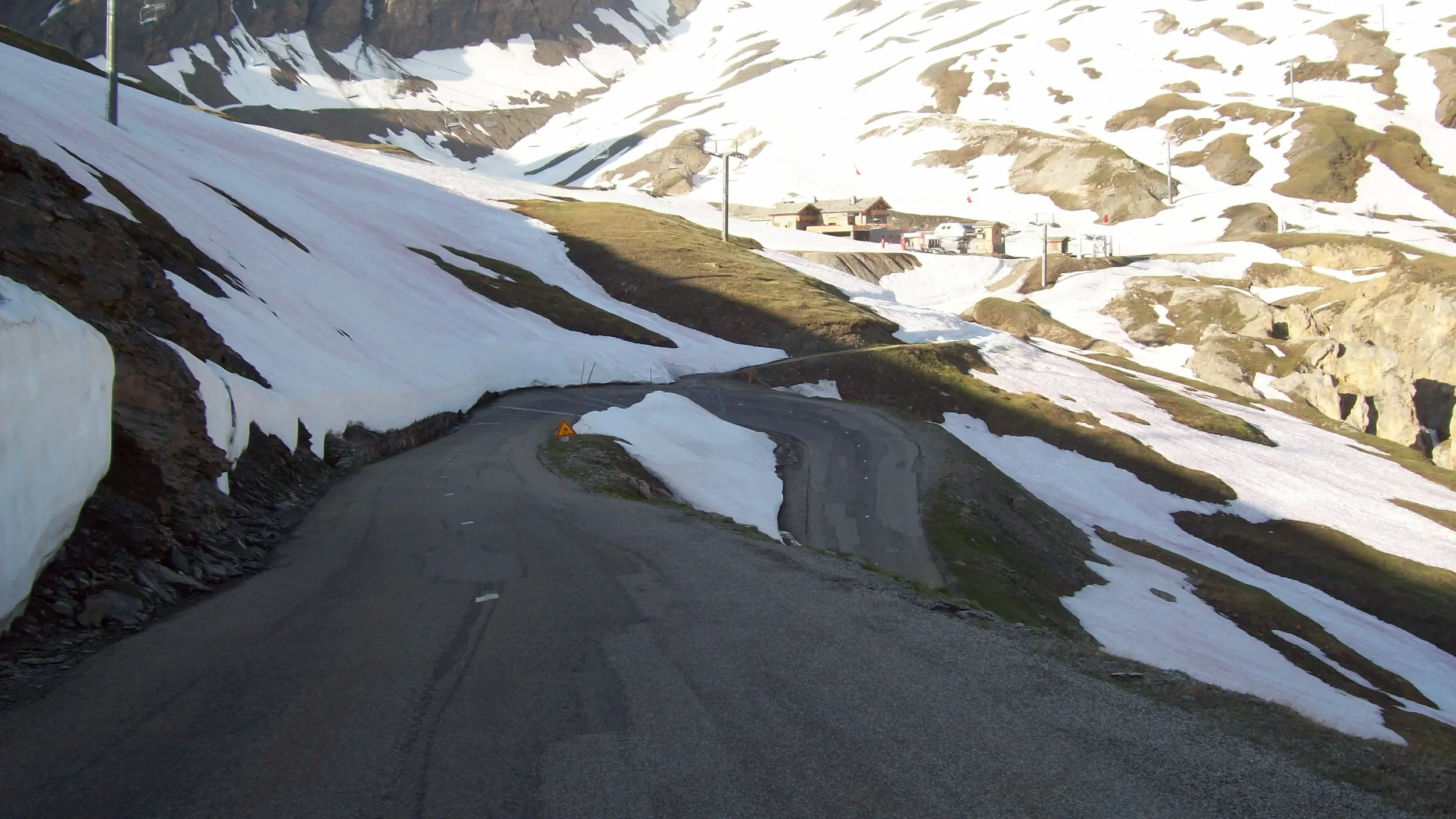 A mountain road winding through partly snow-covered terrain with some exposed rocky patches, leading towards distant buildings and ski lifts on a hillside.