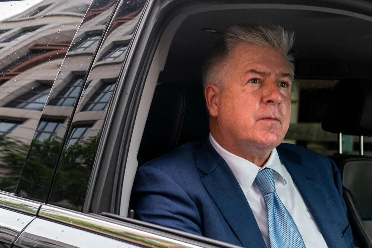 A man with gray hair, dressed in a dark suit, white shirt, and blue tie, is sitting in the backseat of a black car, looking out the window with a serious expression.