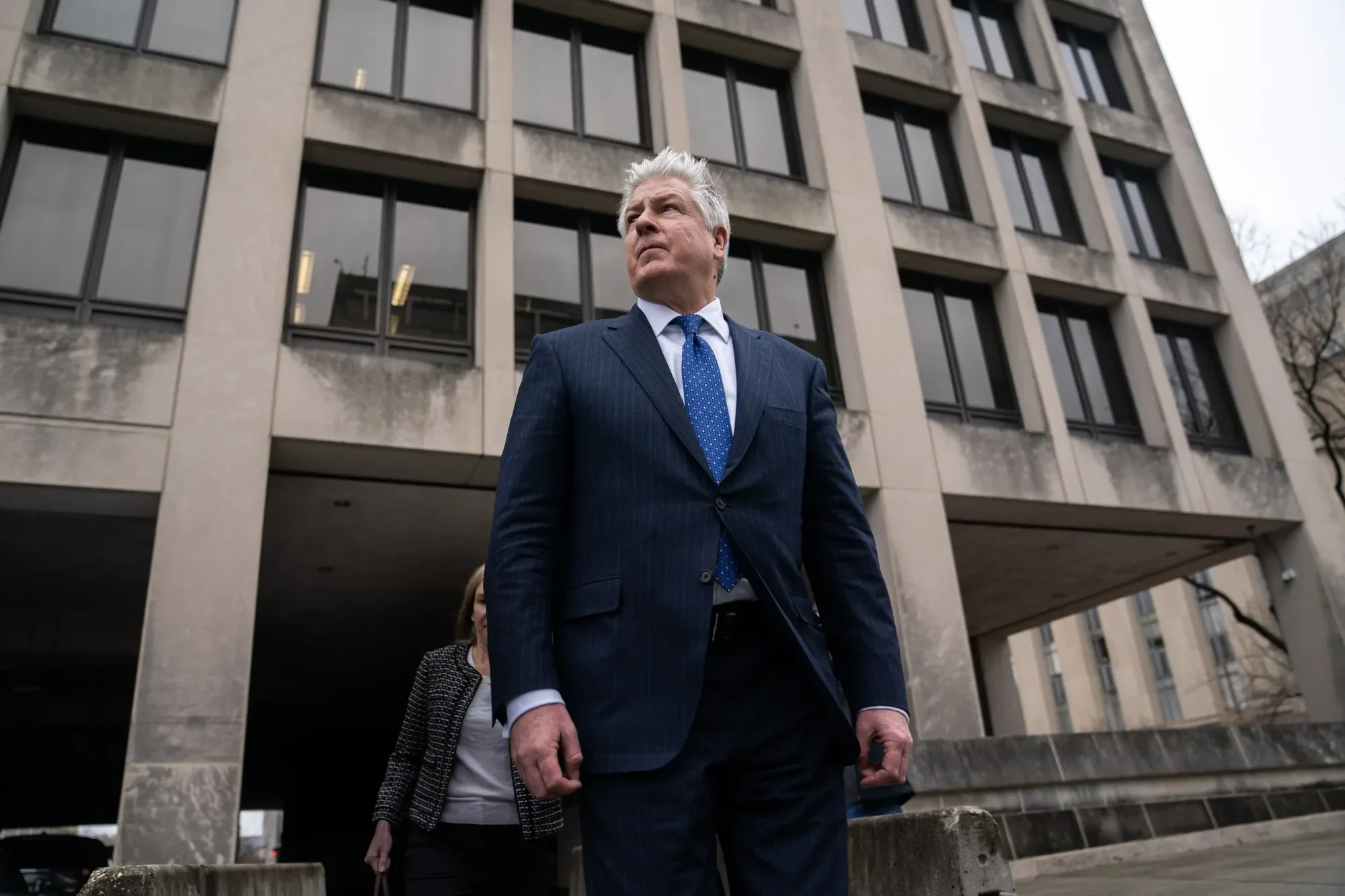 A suited man looking up, standing outside a large concrete building with windows, with a woman walking behind him.