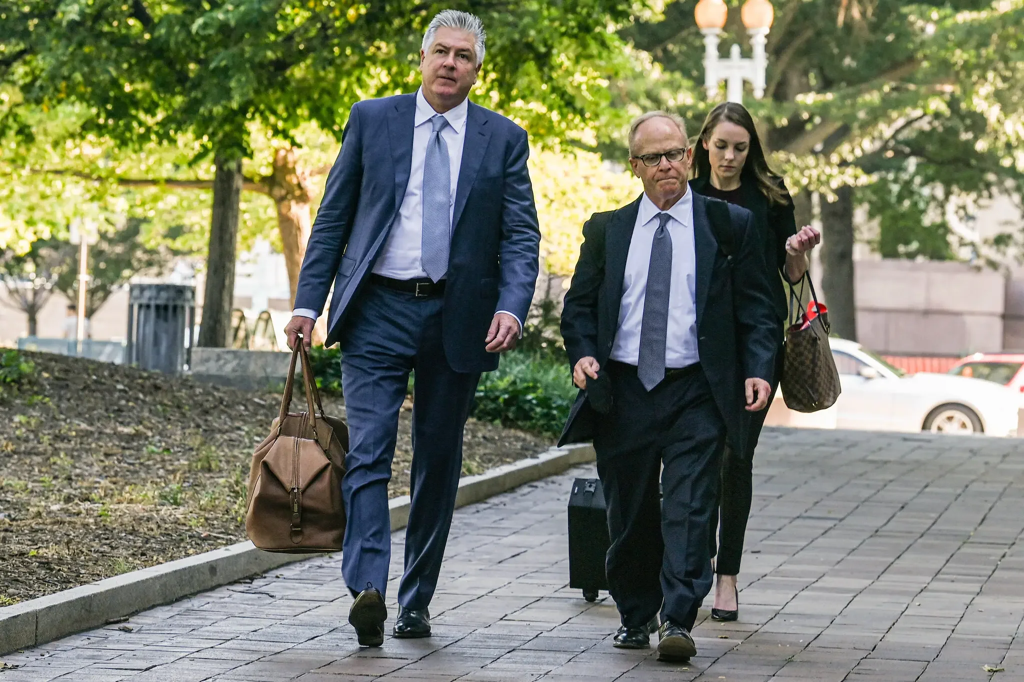 Three professionally dressed individuals walking on a sidewalk with trees and cars in the background, carrying briefcases and handbags.