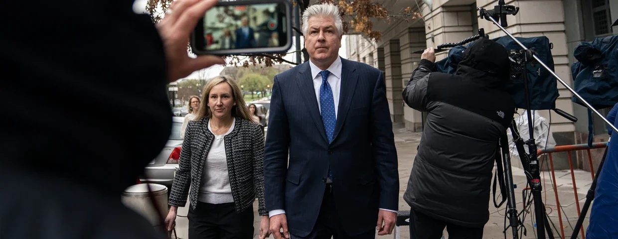 Two individuals, a man and a woman, stand on a city sidewalk during a media event, with a cameraman filming them and a person taking a photo.