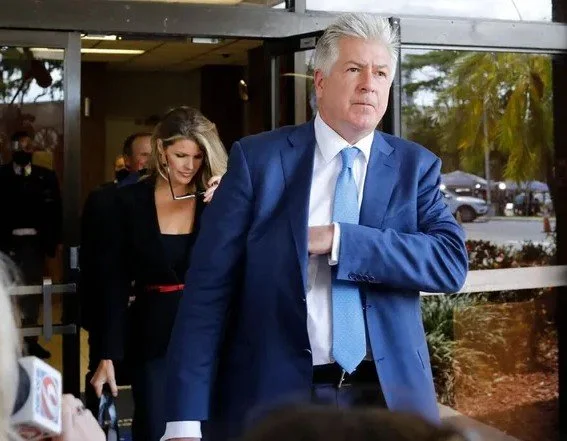 A man in a blue suit and light blue tie with white hair is walking outside, holding his arm across his chest. In the background, a woman with blonde hair wearing a black outfit with a red belt is walking behind him, holding papers, and a few people are visible through the glass windows.