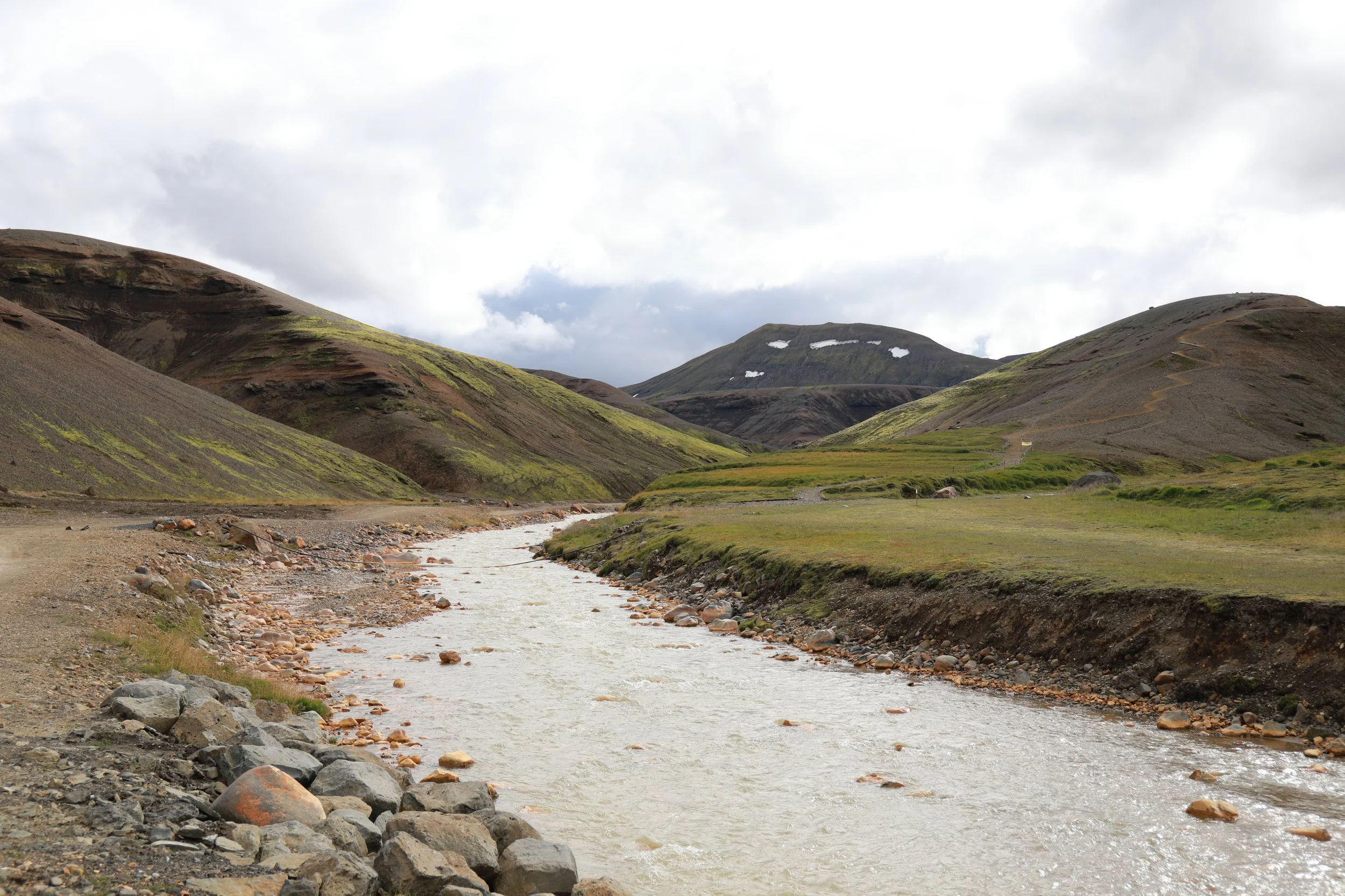 Eine Flusslandschaft in einer bergigen Region mit grünen Hügeln, steinigen Ufern und bewölktem Himmel.