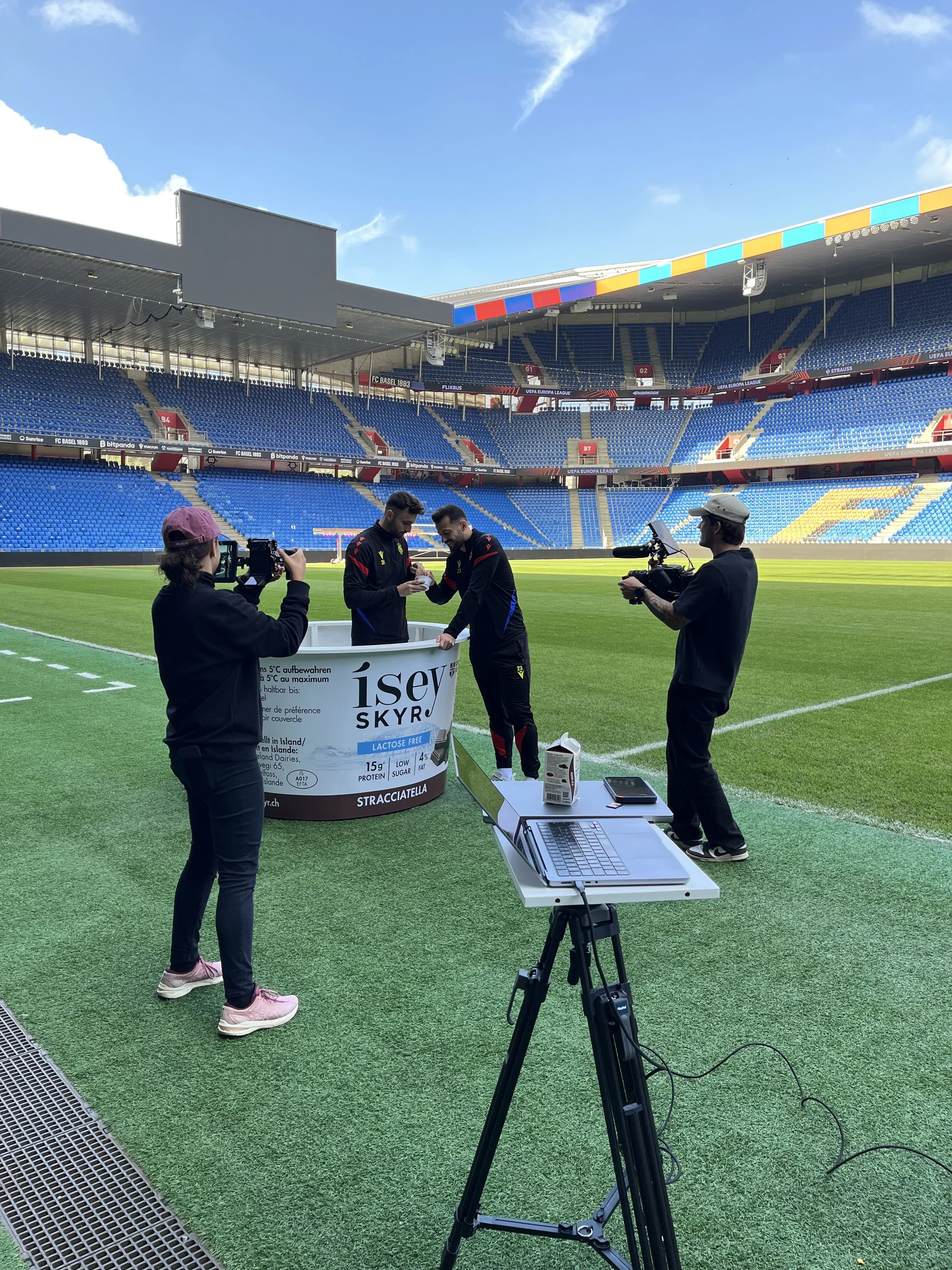 Zwei Männer auf einem Fußballfeld bei einem Interview, umgeben von Kameraleuten, mit einem großen leeren Stadion im Hintergrund.