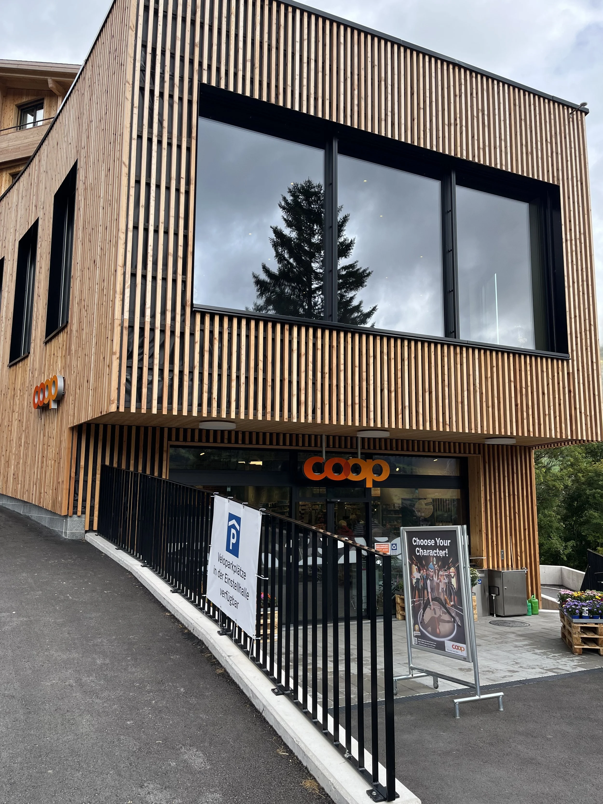Modernes Gebäude mit Holzfassade und großen Fenstern, vorne Eingang mit Coop-Schild, Pegelschilder für Parkplätze und Werbemotiv, Parkrampe links, umgeben von Bäumen und Blumen