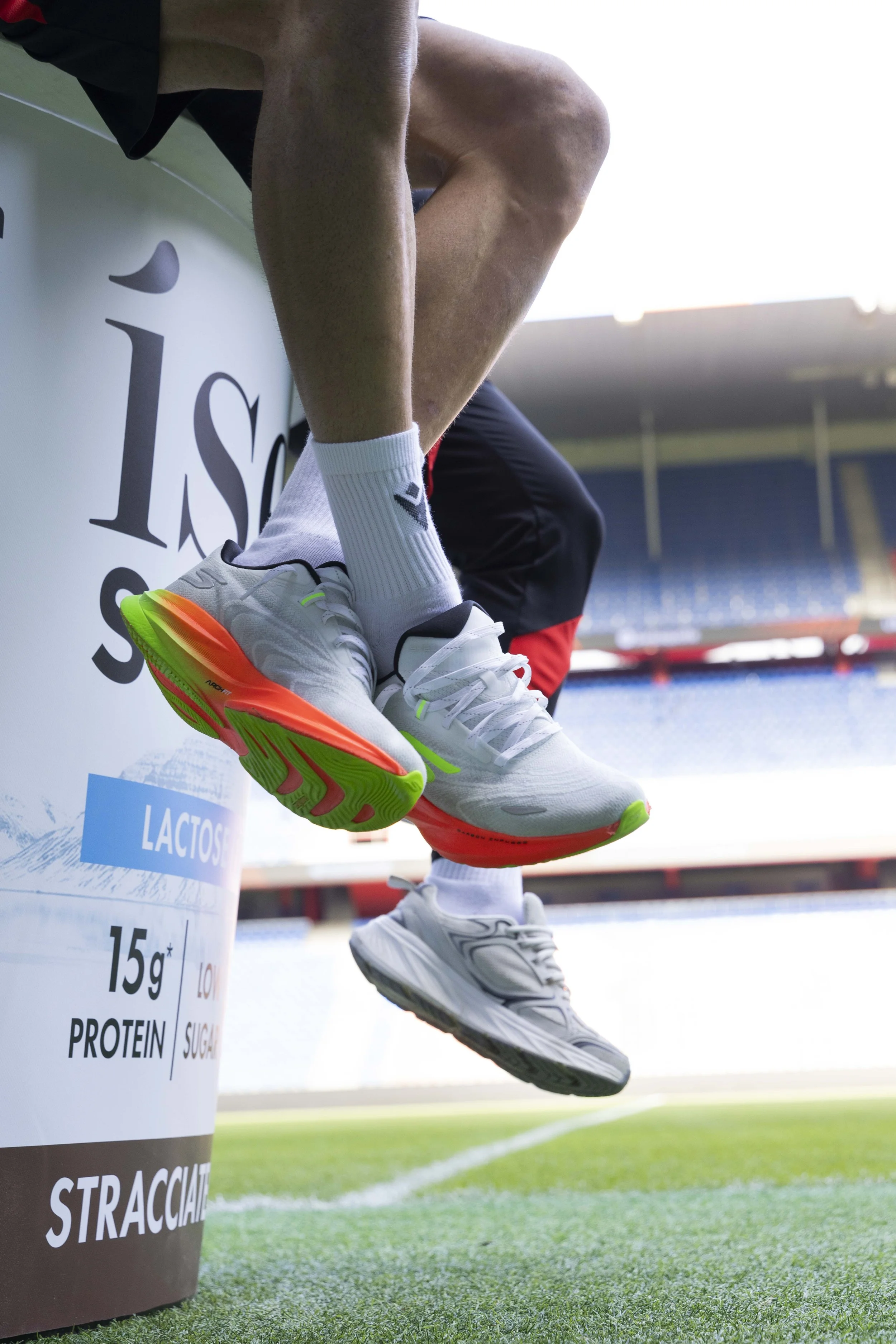 Nahaufnahme eines Spielers, der auf einem Fußballfeld sitzt, mit weißen Turnschuhen und weißen Socken, im Hintergrund ein leeres Stadion.