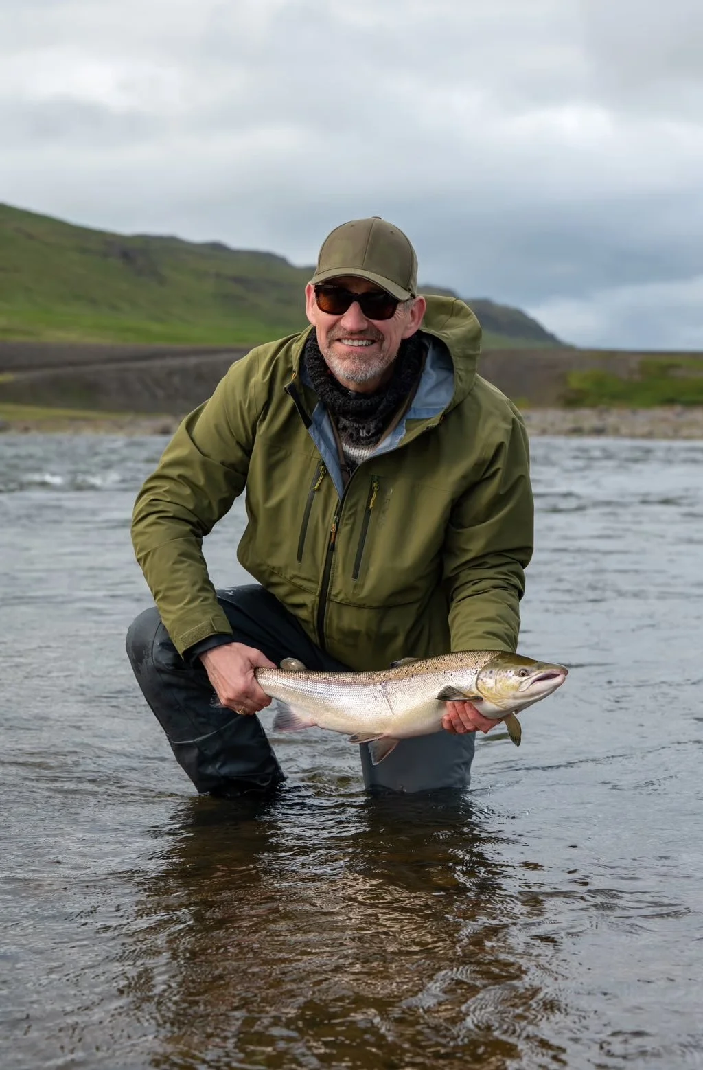 Mann mit Kappe und Sonnenbrille, der einen großen Fisch im Fluss hält, umgeben von einer grünen Hügellandschaft unter bewölktem Himmel.