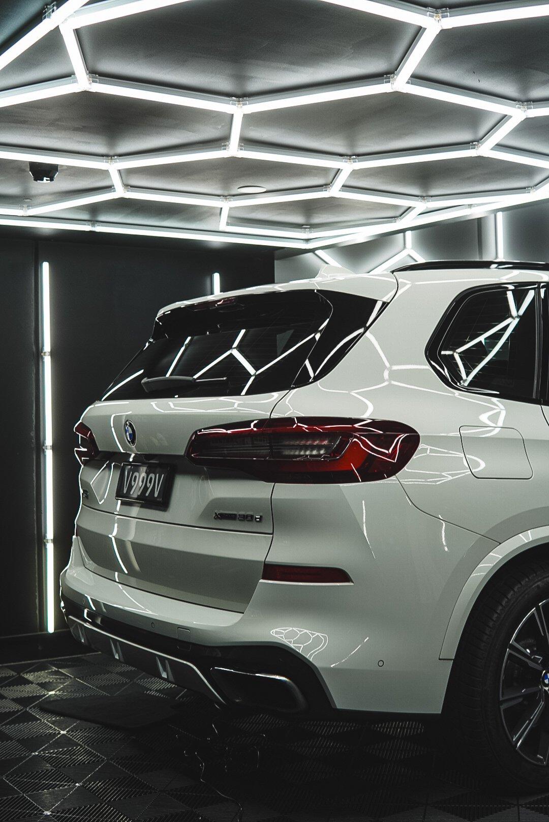 A white BMW SUV parked in a futuristic, dark garage with neon hexagon lighting on the ceiling and vertical light strips on the walls.