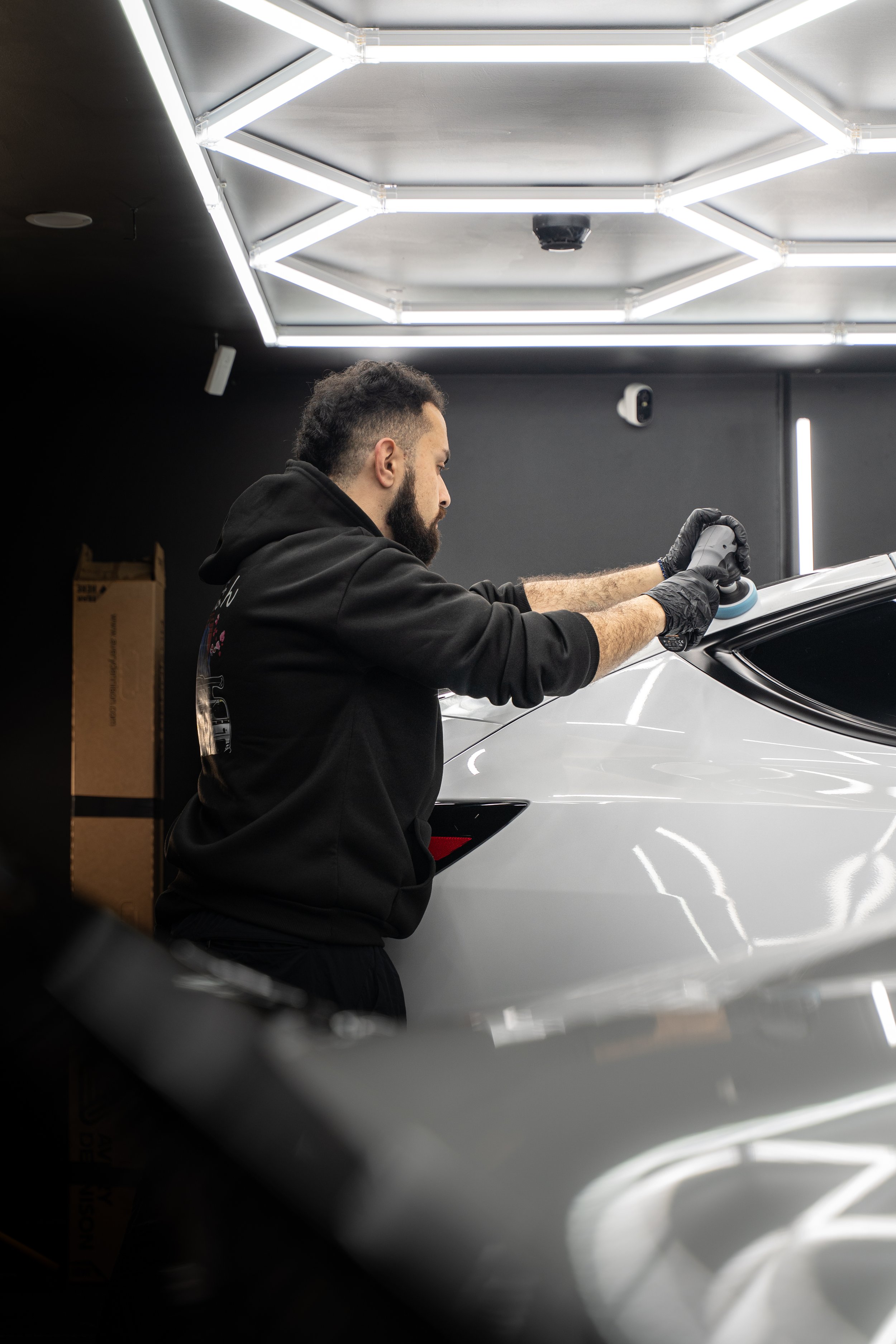 A man wearing black gloves polishing a silver sports car in an automotive workshop with modern lighting.