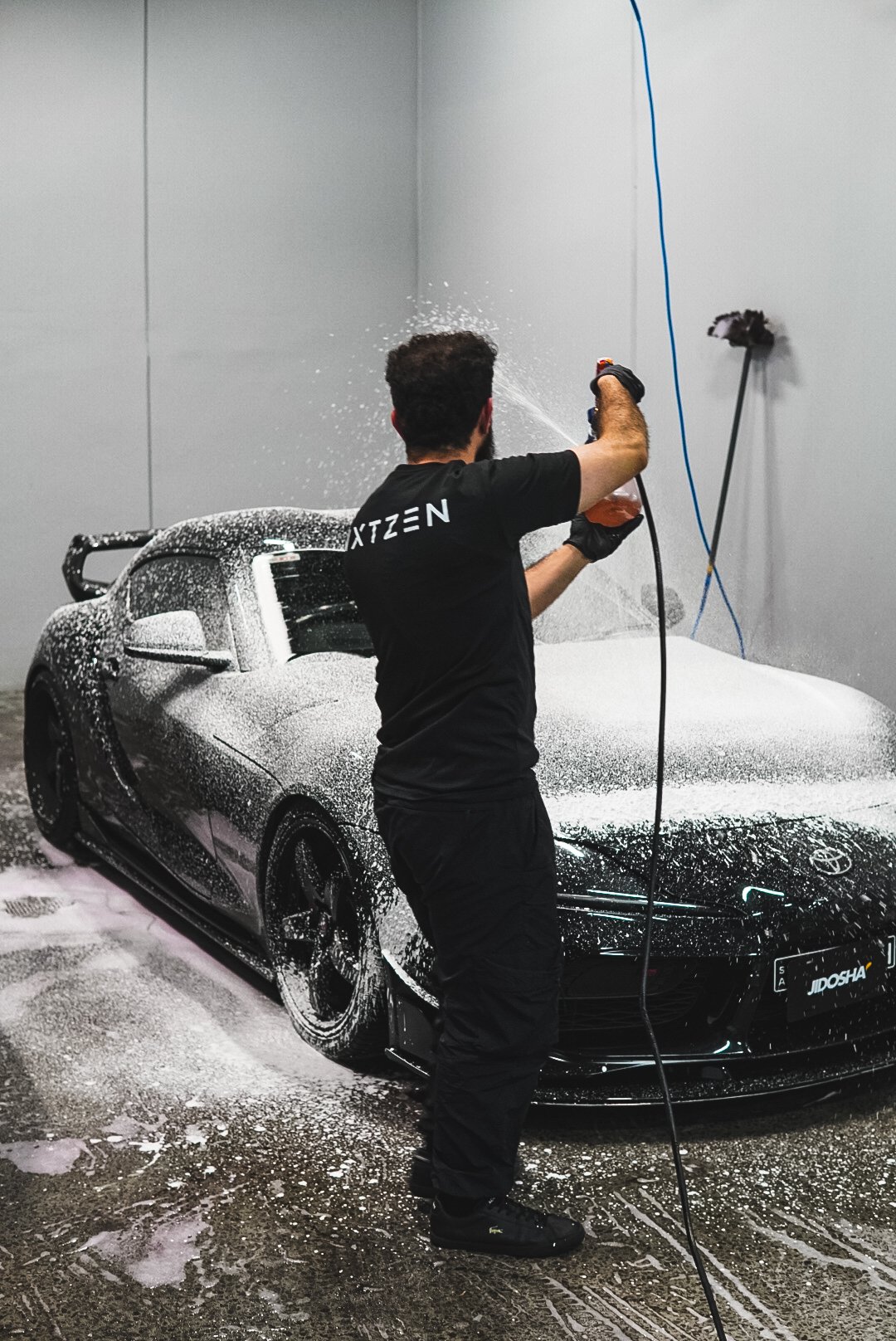 A man washing a black sports car in a car wash station, with foam and water on the vehicle and the floor.