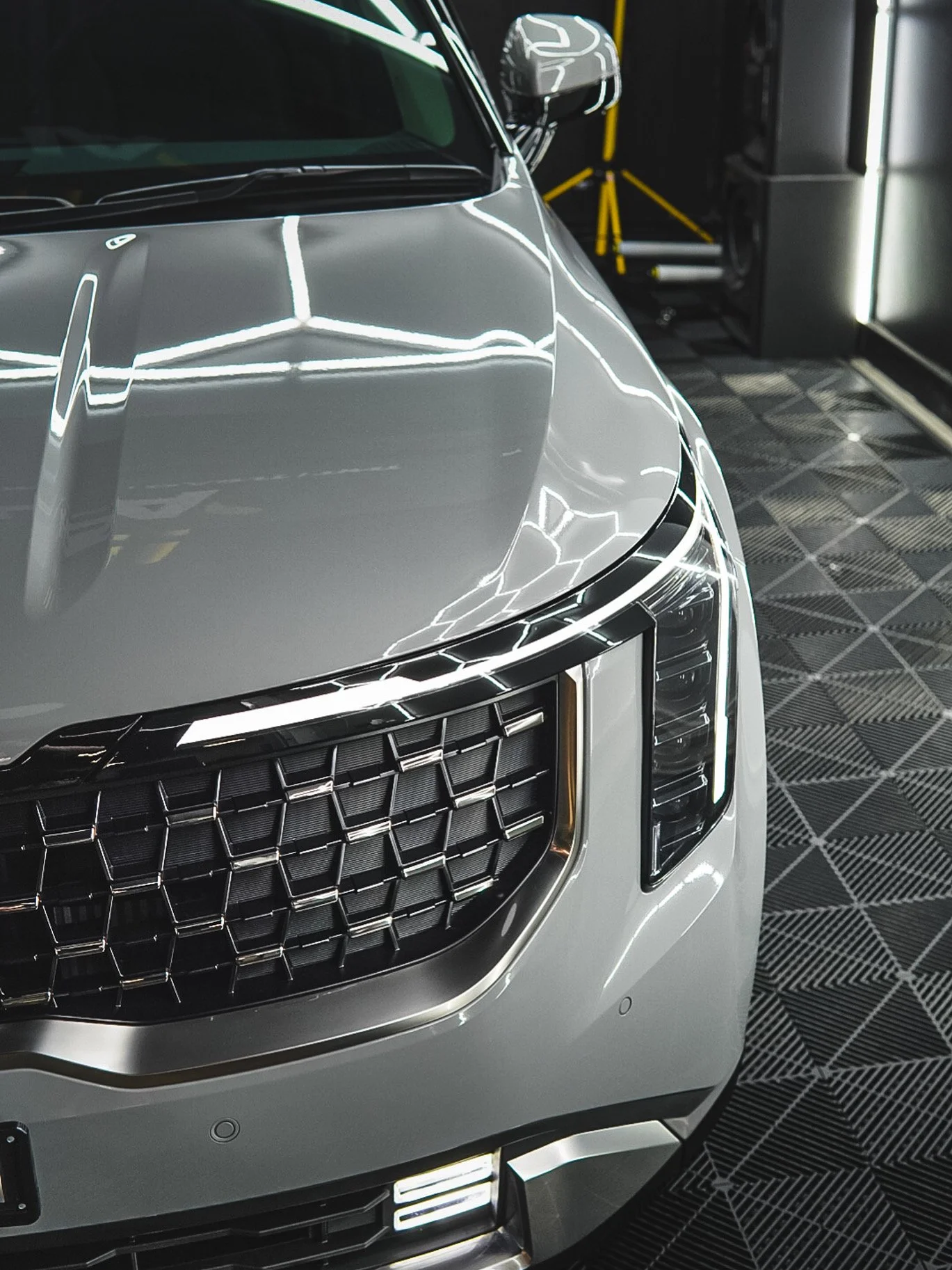 Close-up of the front end of a silver luxury SUV parked indoors, showing the grille, headlights, and reflection of bright ceiling lights.