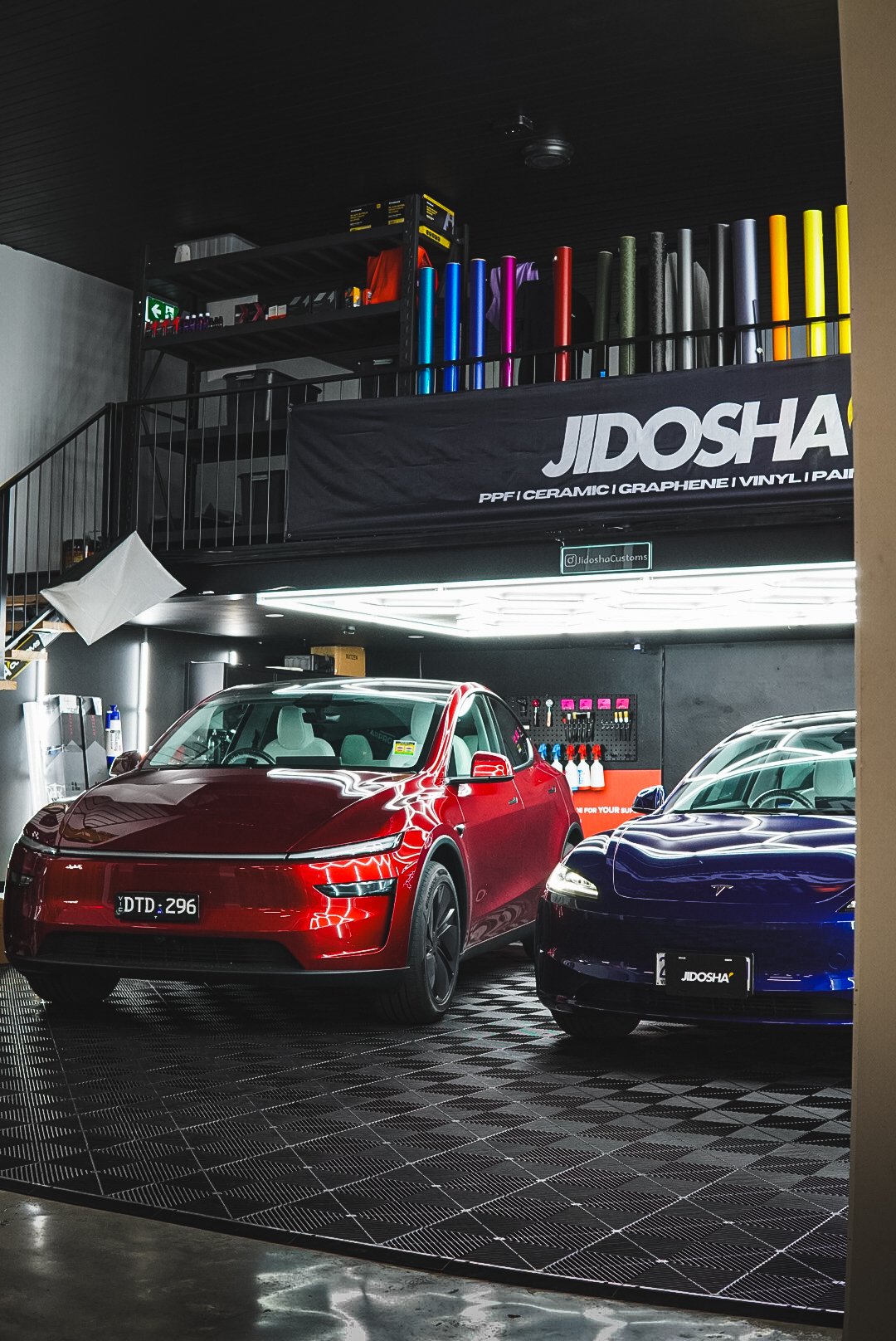 Two Tesla cars, one red and one dark, parked inside a automotive wrapping shop with various colorful vinyl wraps stored on a mezzanine level.