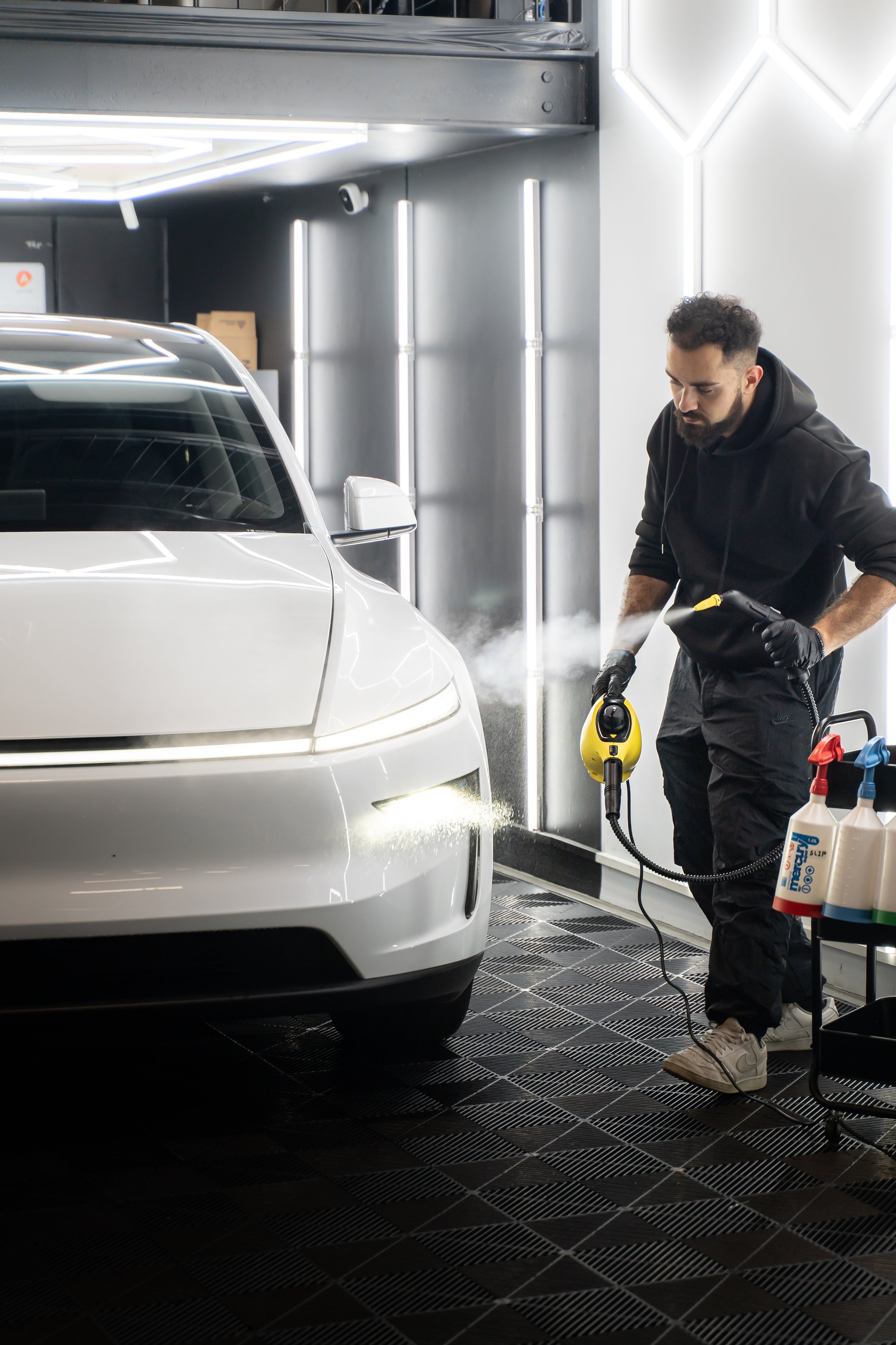 A man in a black hoodie and gloves cleaning a white car with a steam cleaner in a modern, well-lit garage.