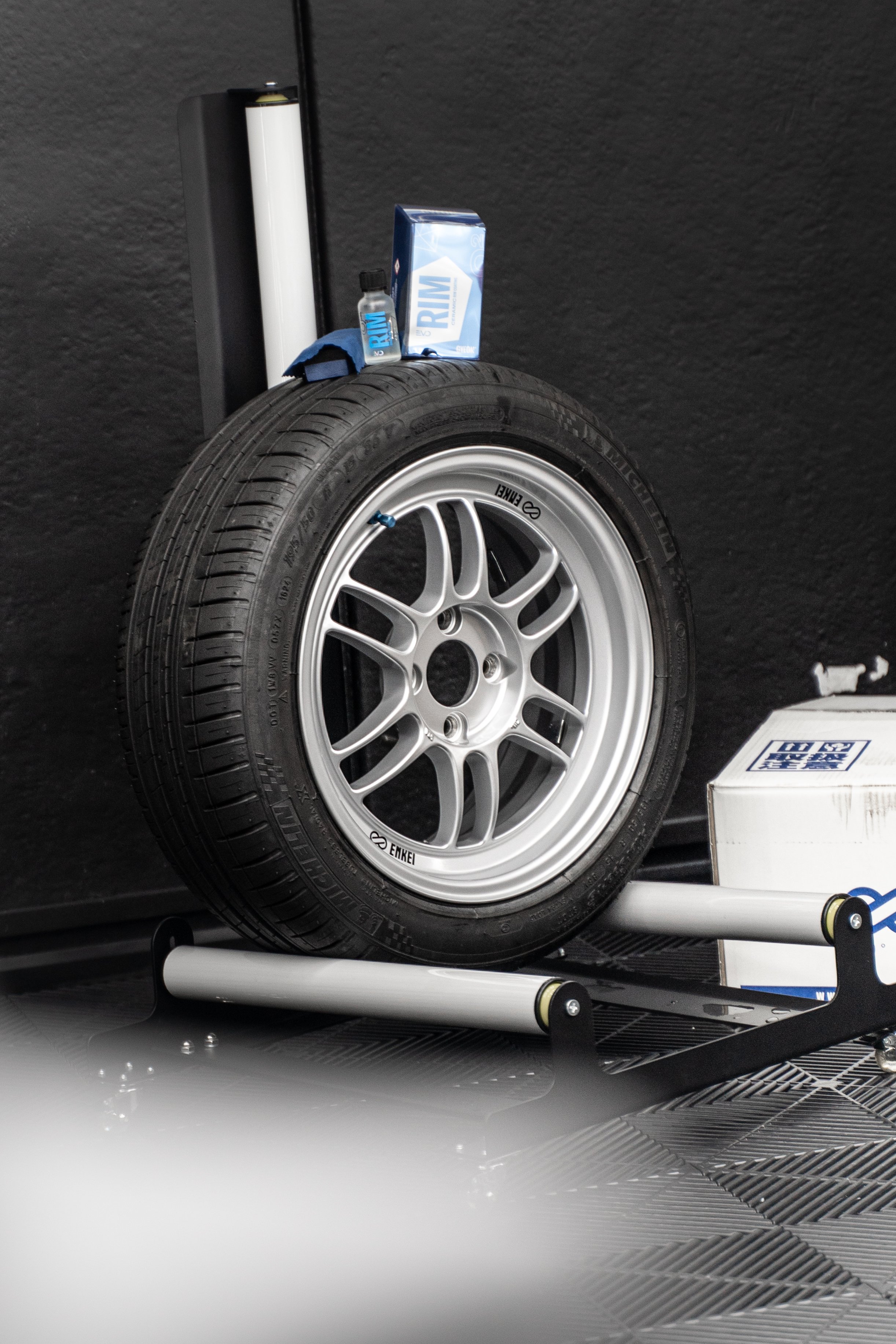 A racing tire mounted on a wheel on a wheel balancing machine, with a box, a small bottle, and a box of RIM cleaner on a shelf in the background.