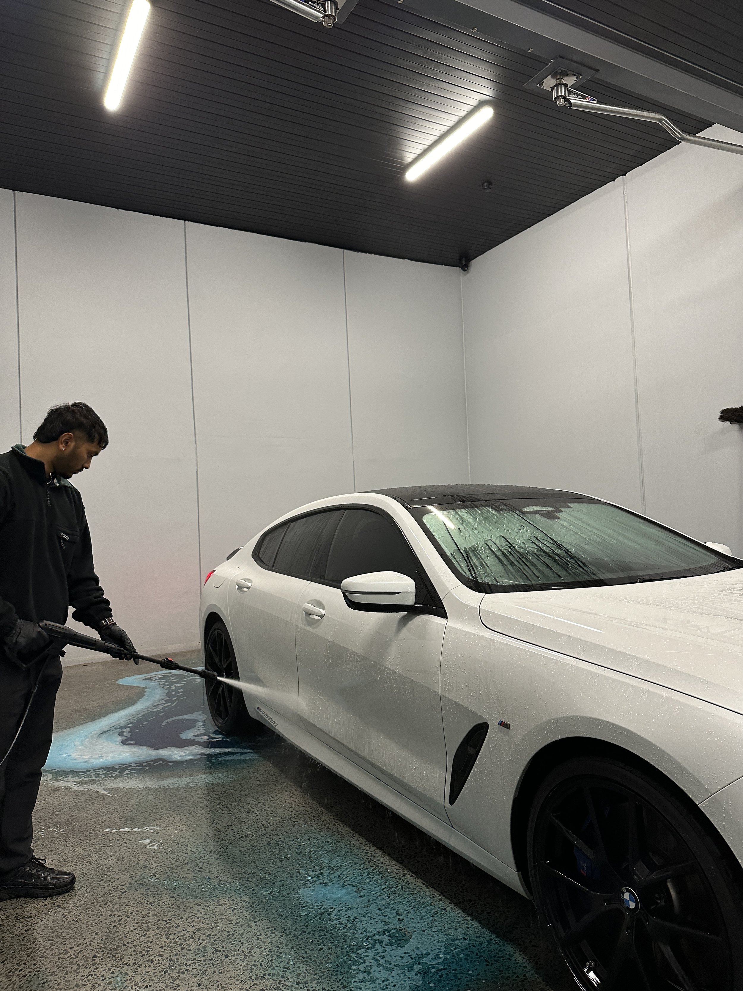 A person washing a white BMW car inside a car wash bay with dark ceiling and white walls.
