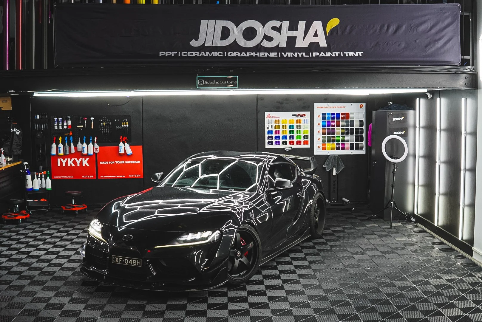A black sports car inside a well-lit garage with a black wall, decorated with posters for car care products, paint colors, and brand logos, and a black canopy at Jidosha Melbourne
