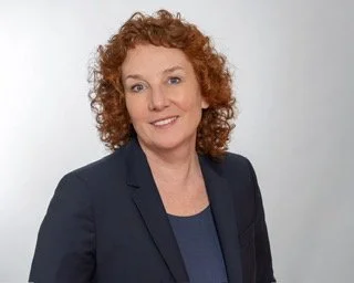 Porträt einer Frau mit roten, lockigen Haaren in dunkler Blazerjacke auf weißem Hintergrund.