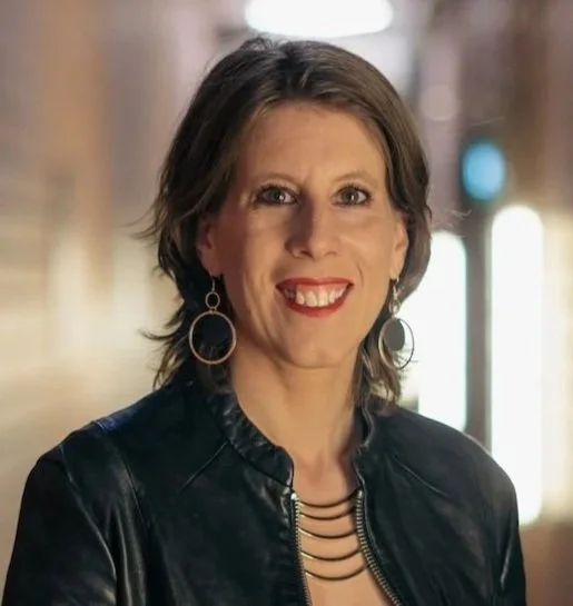 Eine Frau mit braunen, schulterlangen Haaren, rotem Lippenstift und Ohrringen, die eine schwarze Lederjacke trägt, steht in einem Stadtumfeld bei Nacht.