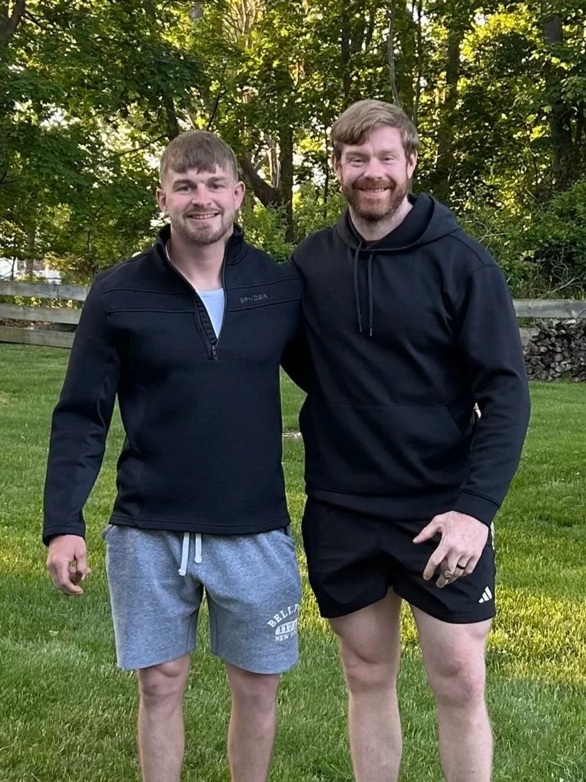 Two men standing outside on a grassy area, smiling, with trees and a wooden fence in the background.