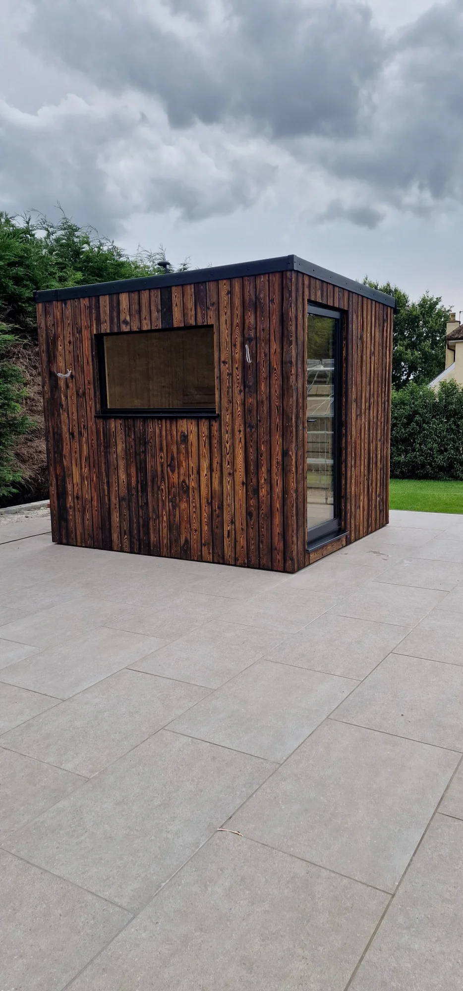 Small modern wooden structure with large window and glass door, situated on a tiled outdoor patio under cloudy sky.