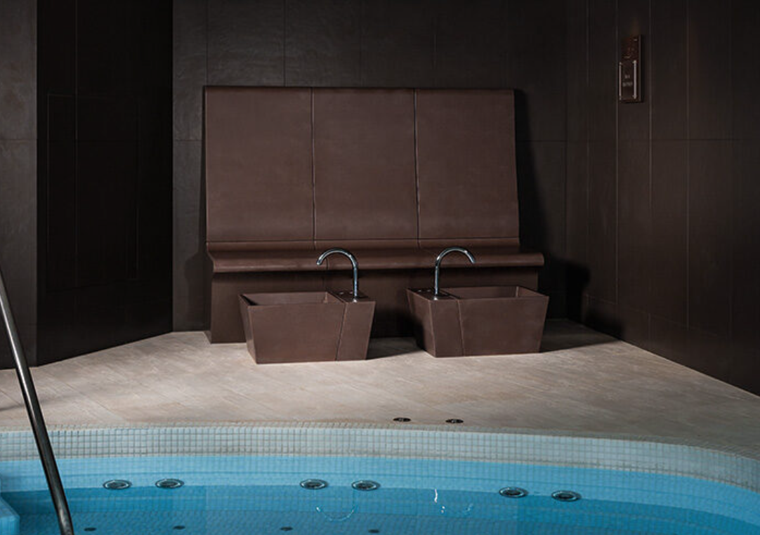 Indoor spa area with two foot baths, brown leather bench, and a small portion of a swimming pool with blue tiles.