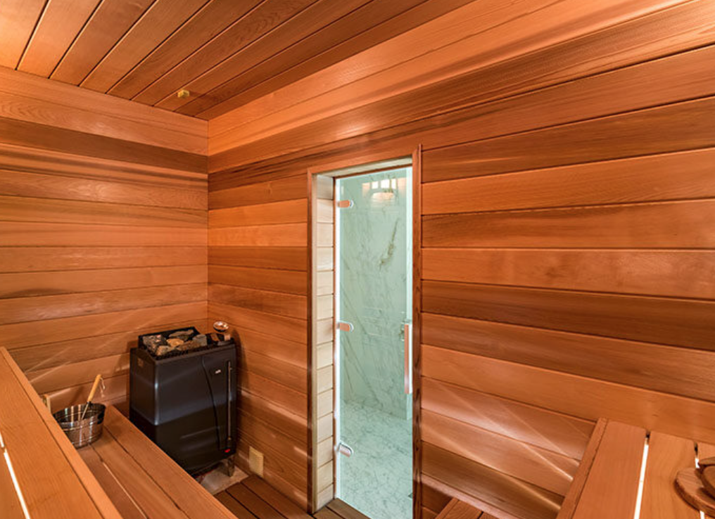 A sauna room with wooden walls, a glass door to a shower area, a heater with stones, and wooden benches.