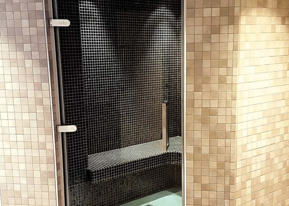 A glass shower door with a stainless steel handle, against a wall of small black tiles, with some beige tiles visible on the adjacent wall.