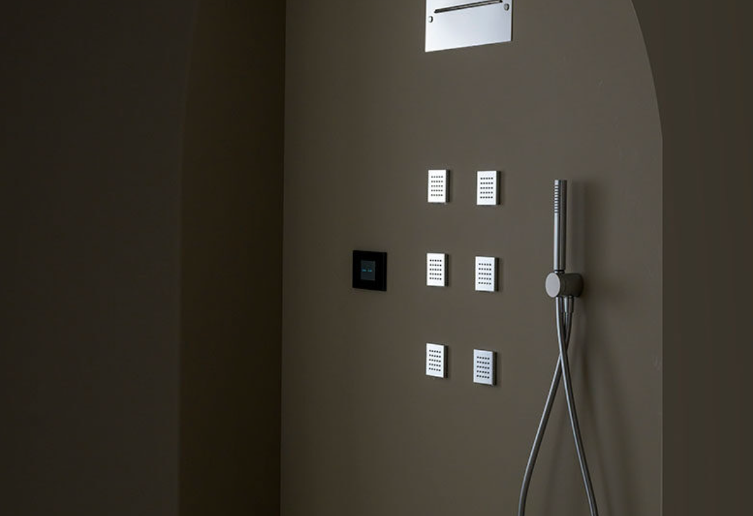 A modern shower setup with a handheld showerhead, multiple control panels, and a small white panel on a beige wall.