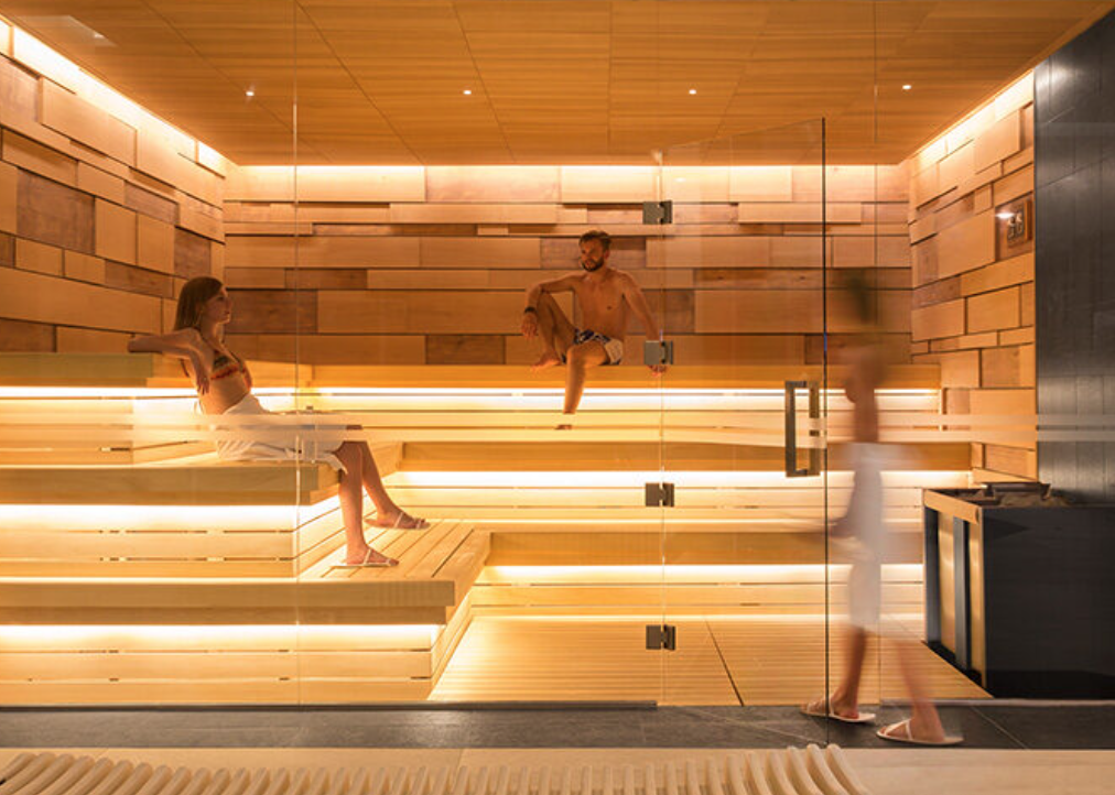 People relaxing in a modern sauna with wooden benches, warm lighting, and glass walls.