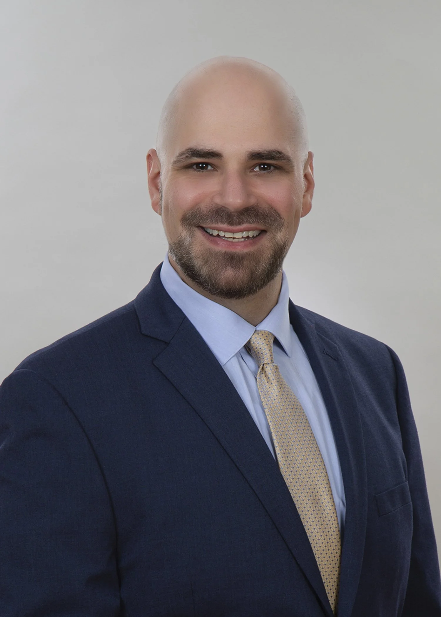 A professional headshot of a smiling bald man with a beard, wearing a navy suit, light blue shirt, and a yellow patterned tie against a plain light background.