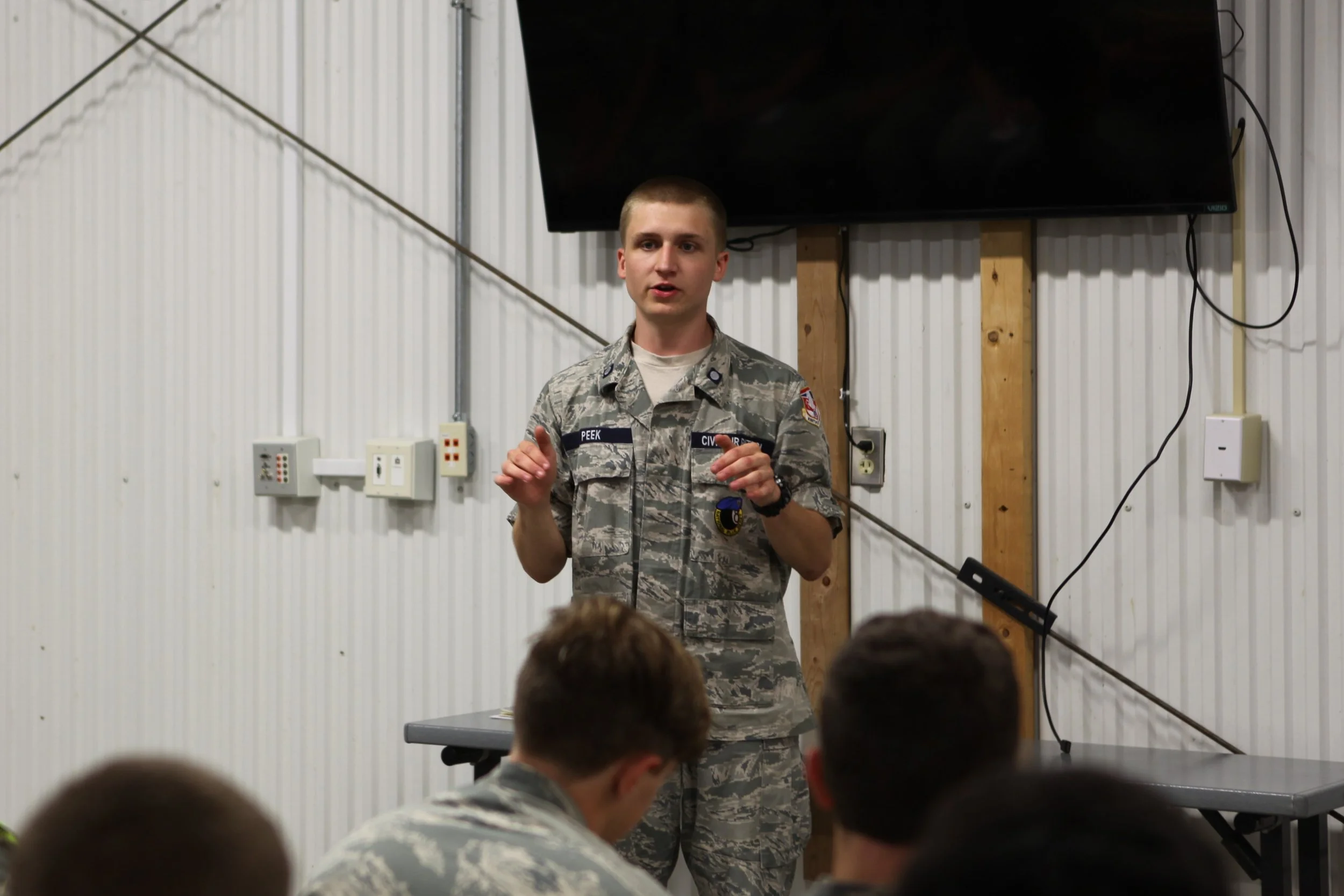 A young man in military uniform giving a presentation to a group of soldiers in an industrial room with white corrugated metal walls and a large flat screen TV.