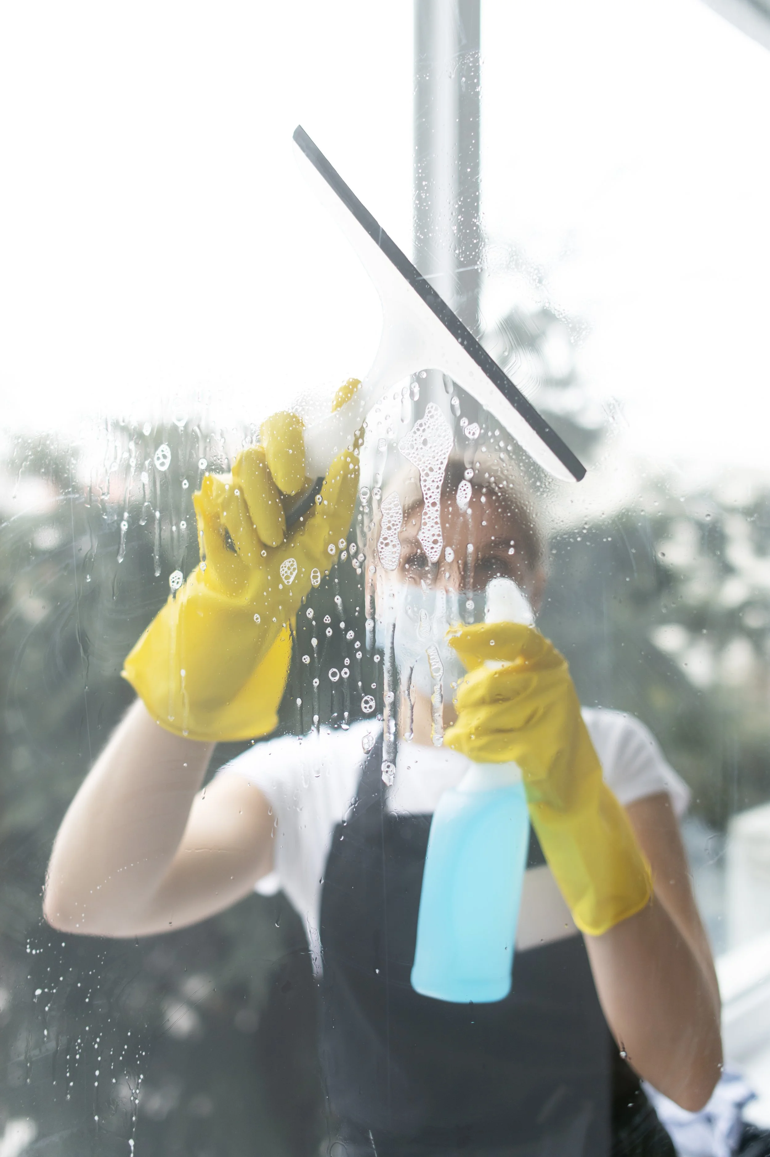 Person cleaning a glass window with a squeegee and spray bottle, wearing yellow gloves, in a bright setting