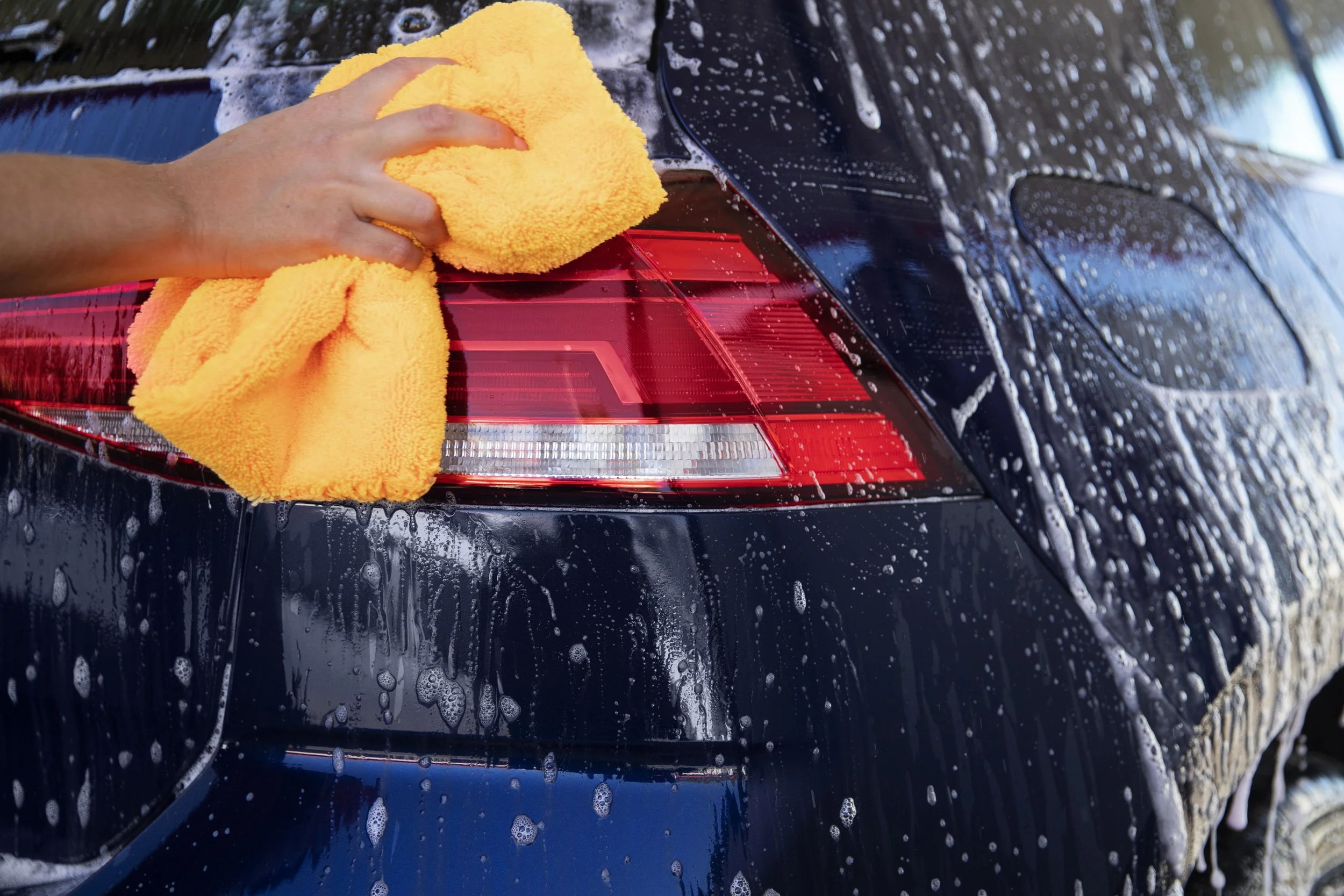Person hand cleaning the rear taillight of a black car with a yellow microfiber cloth during a wash.
