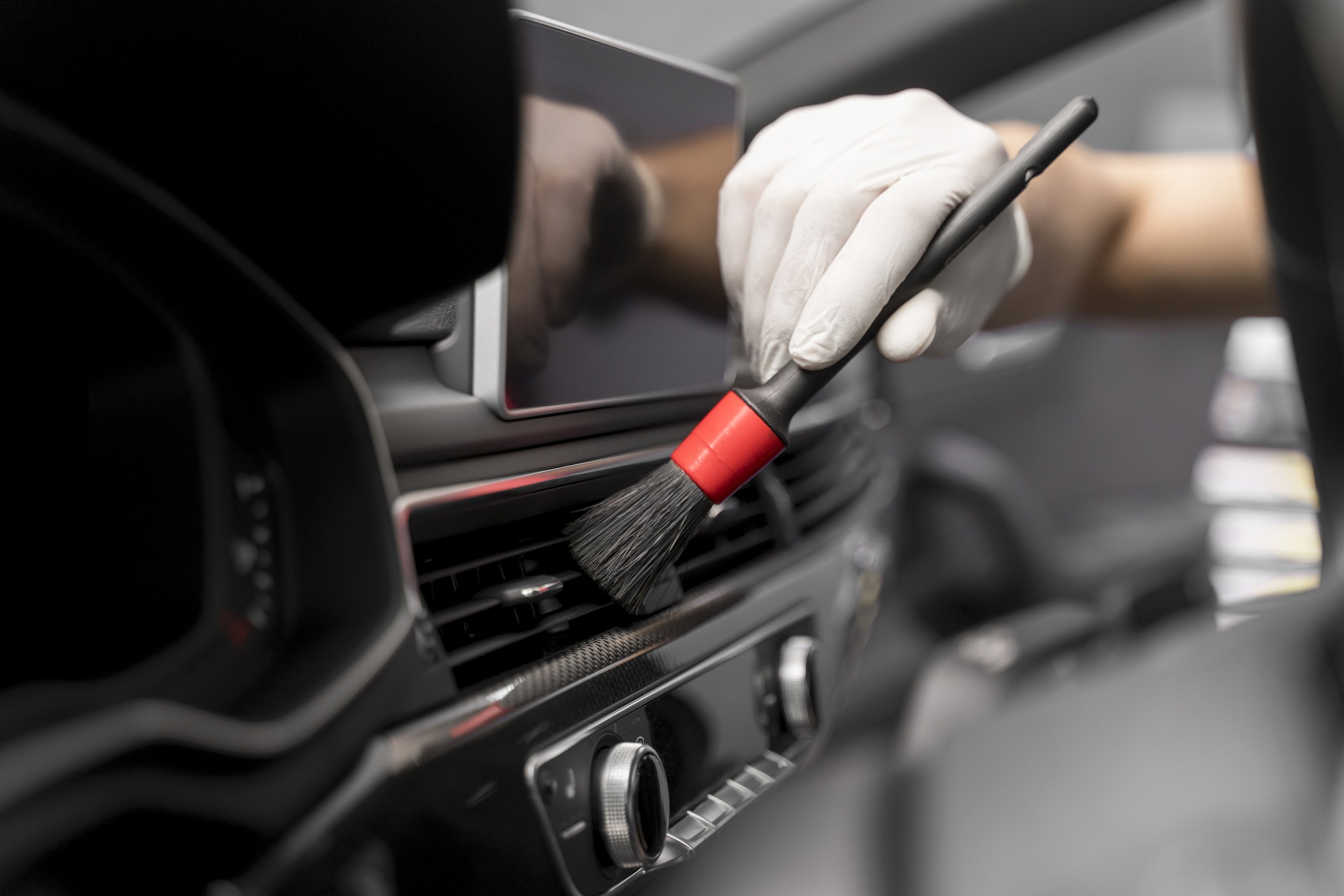Person in white gloves using a brush to clean the air vent of a car dashboard.