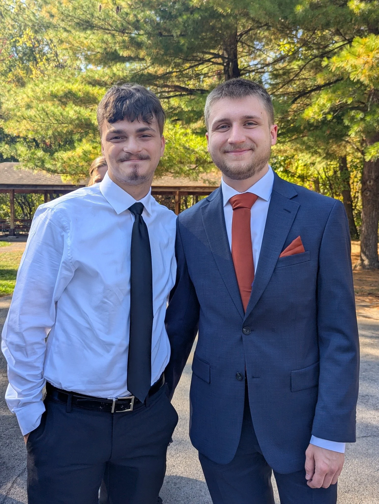 Two young men dressed in formal attire standing outdoors in front of trees, smiling at the camera.