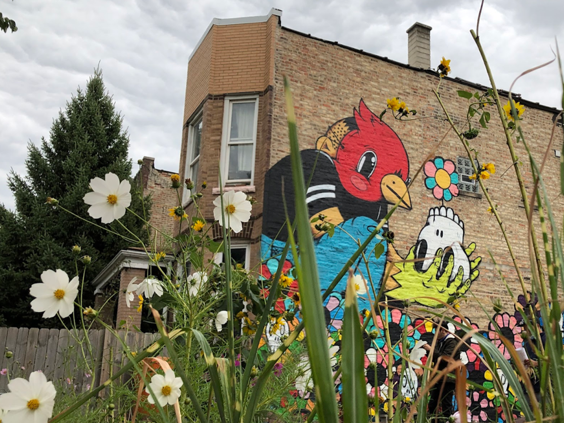 In the foreground, native flowers and grasses partially obscure a mural depicting a boy in a bird mask holding a skull, with a single flower growing out of it.